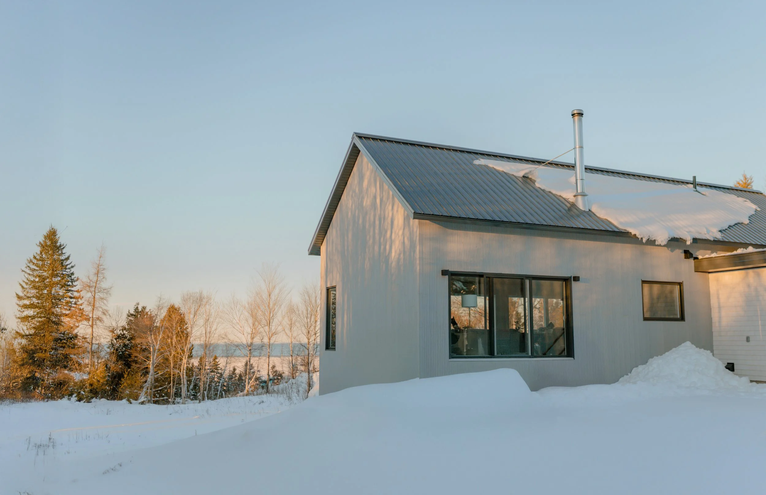 Maison moderne en bois blanc avec toit en métal sous la neige, arbres en arrière-plan, ciel clair.