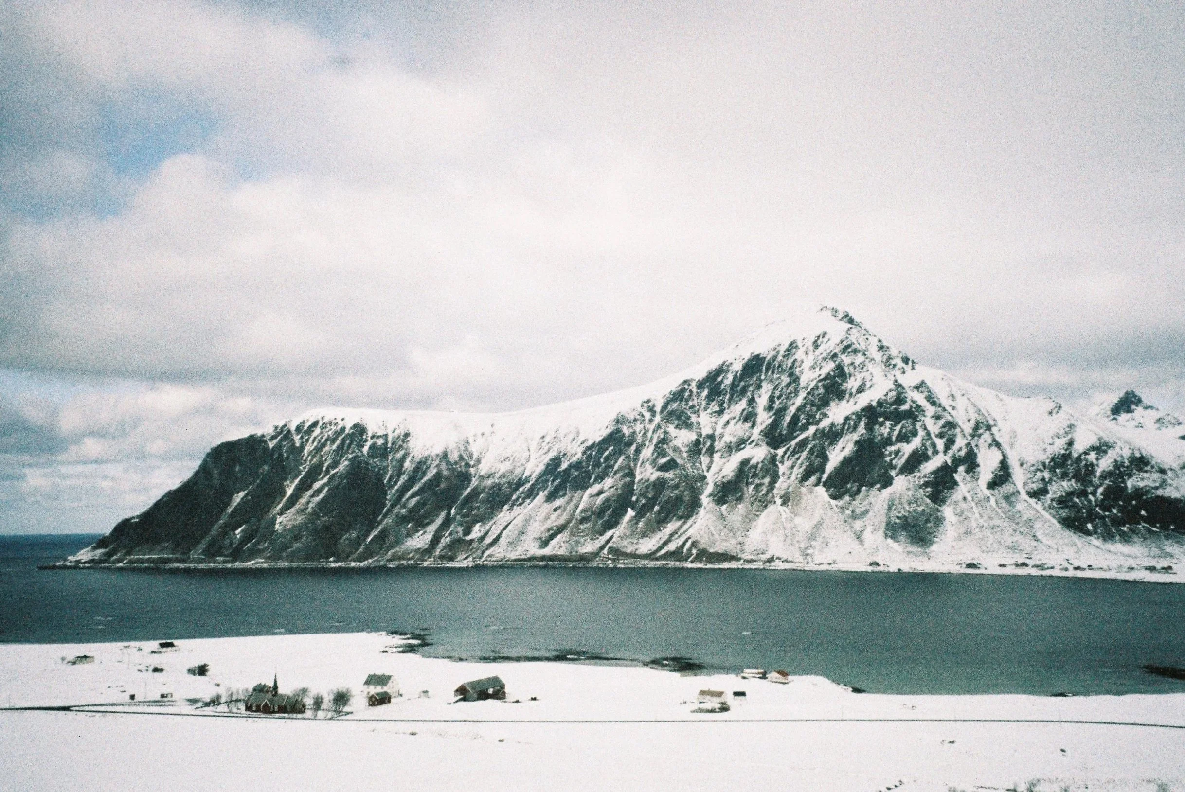 Snow-covered mountain overlooking a body of water with a few small buildings along the shore.
