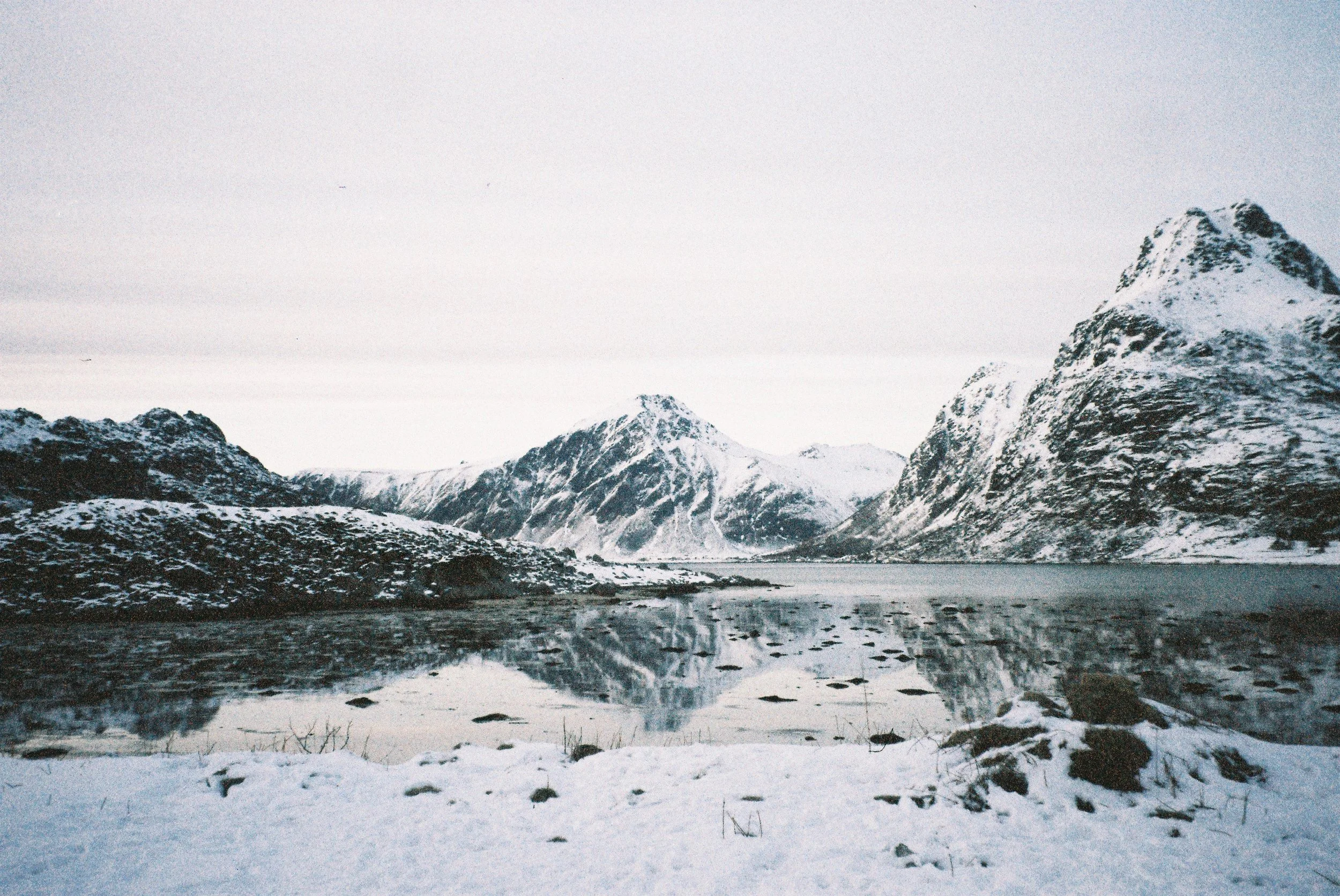Snow-covered mountains surrounding a calm lake reflecting the peaks under a cloudy sky.