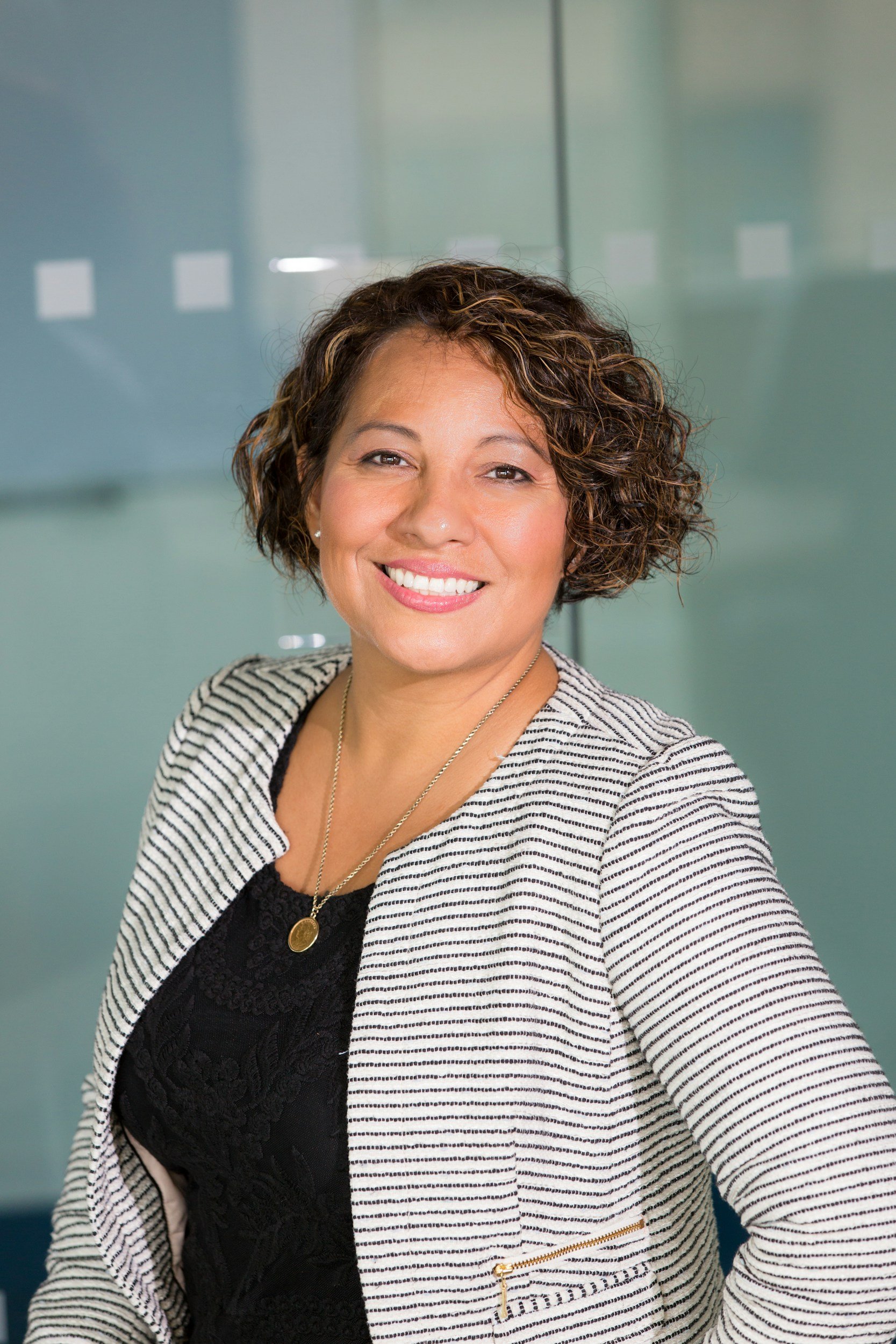 A smiling woman with curly short hair, wearing a black top, a striped blazer, and gold jewelry, standing in front of a glass wall.