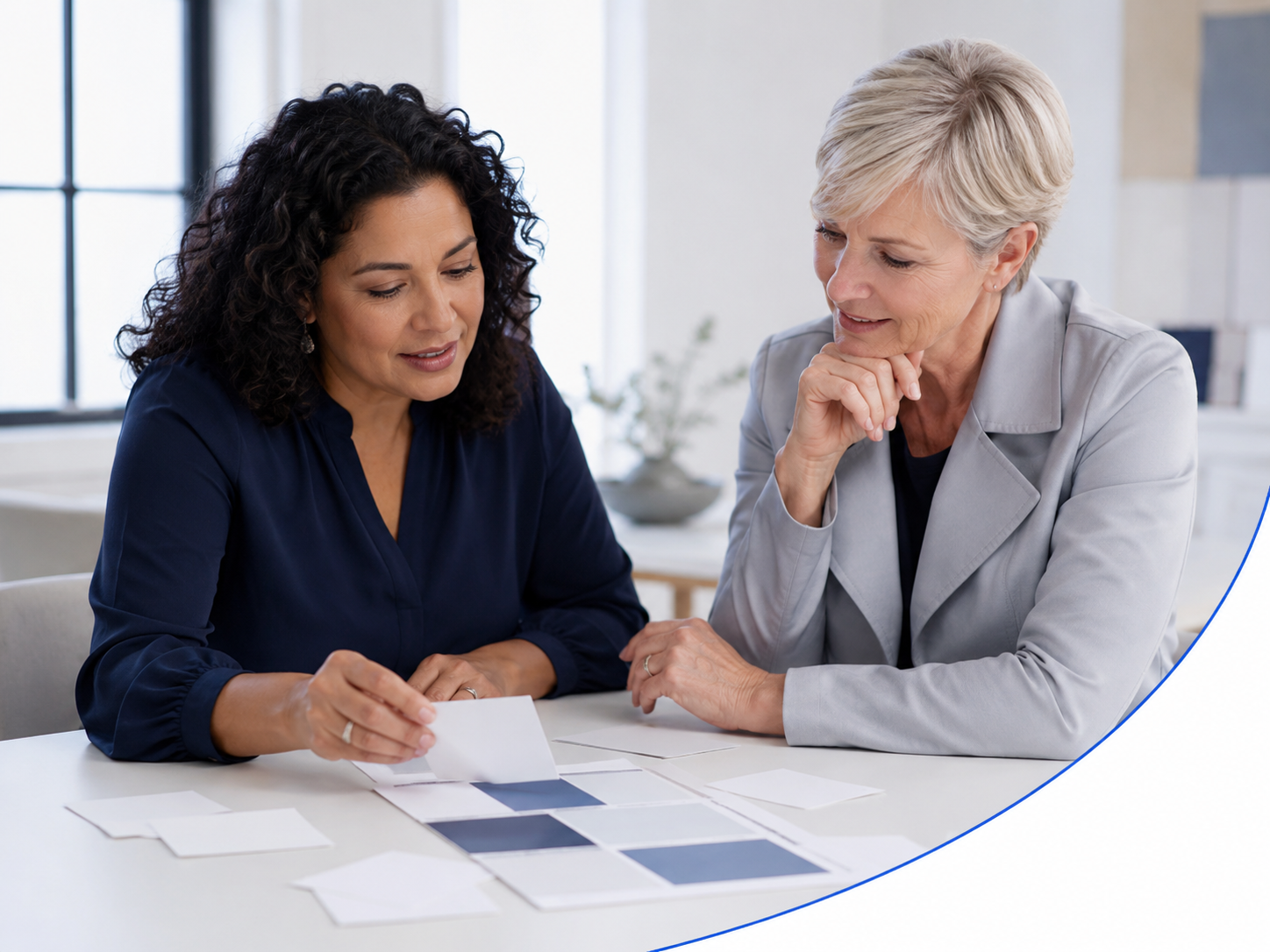 Two women, one with dark, curly hair and the other with short, gray hair, are sitting at a table examining color swatches and paper samples, engaging in a discussion about design or color choices.