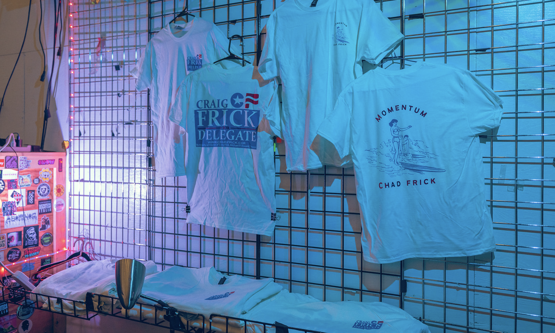 T-shirts hanging on a wire grid display, with campaign slogans and graphics supporting Craig Frick for delegate, illuminated by colorful lighting.