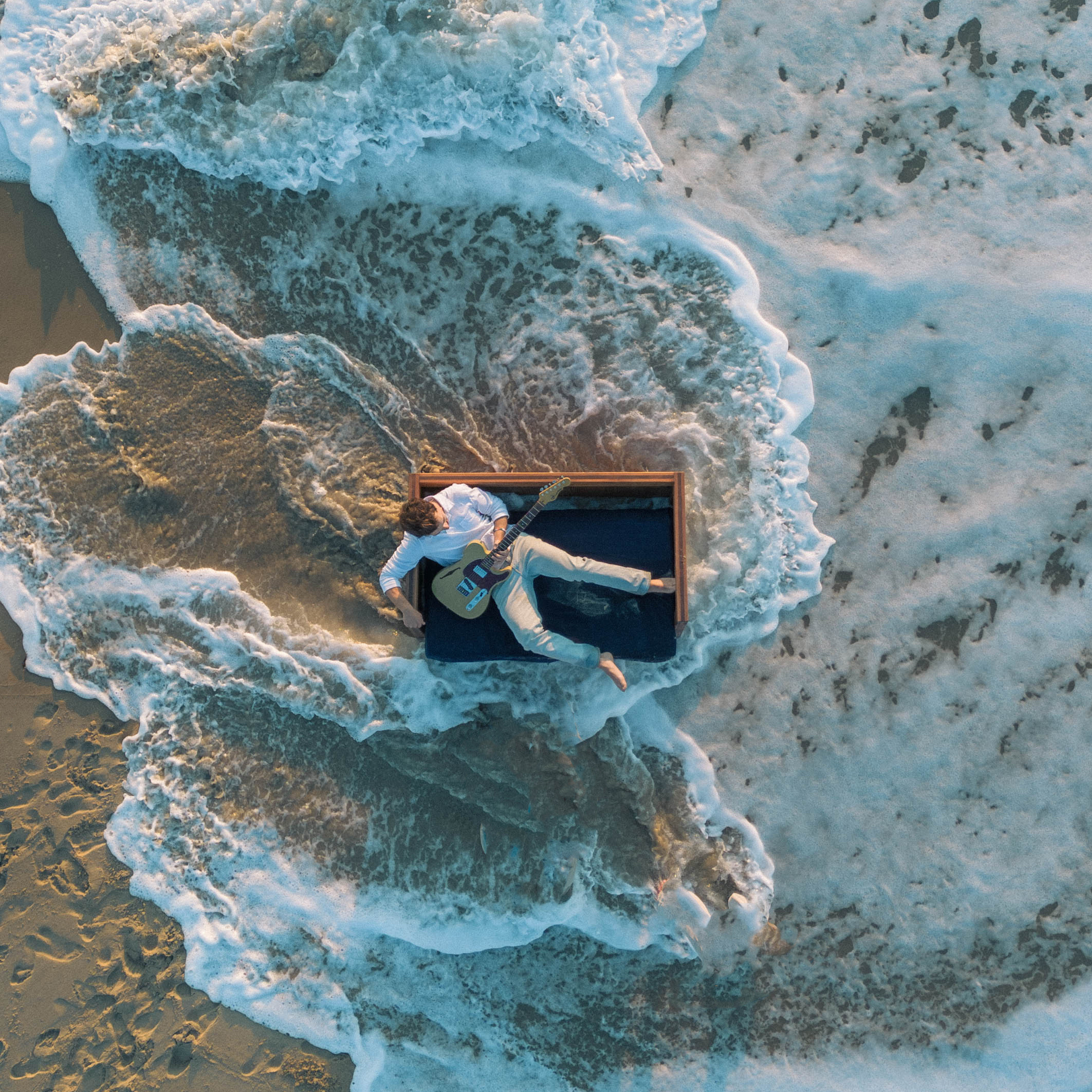 Overhead view of a person playing an electric guitar on the beach, with waves crashing onto the shore.