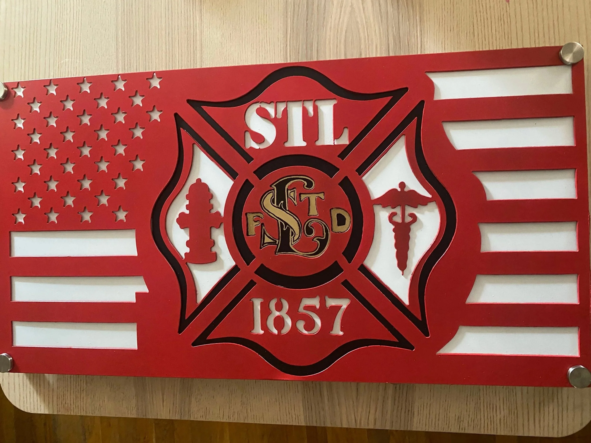 A decorative metal sign with a firefighter emblem, featuring the Maltese cross, an American flag on the left side, and the number 1857 at the bottom.