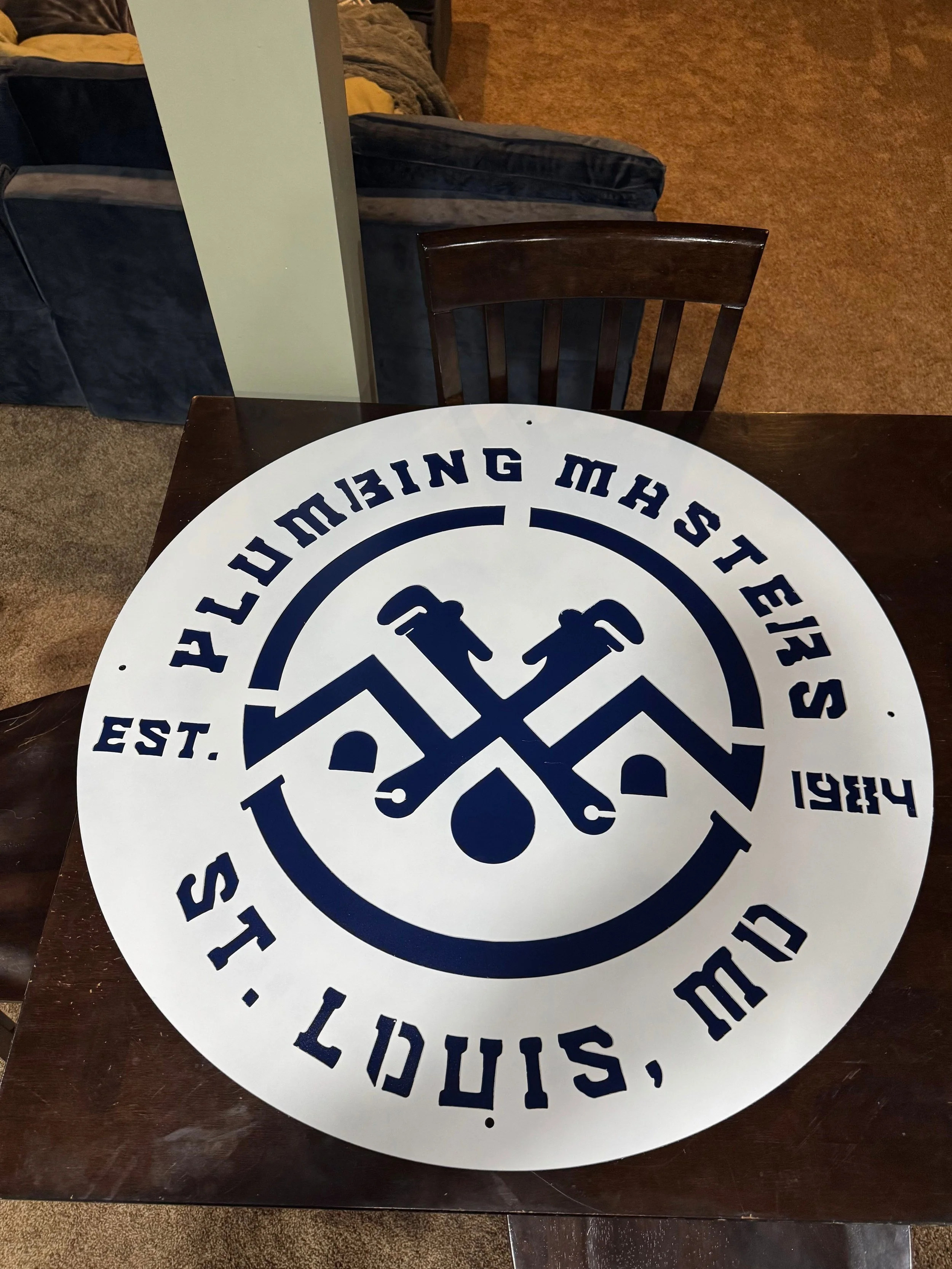 Circular sign with crossed hammer and paintbrush icons at the center, surrounded by the words "Ylumbing Masters," "St. Louis, MO," "Est. 1941," and "Who We Are".