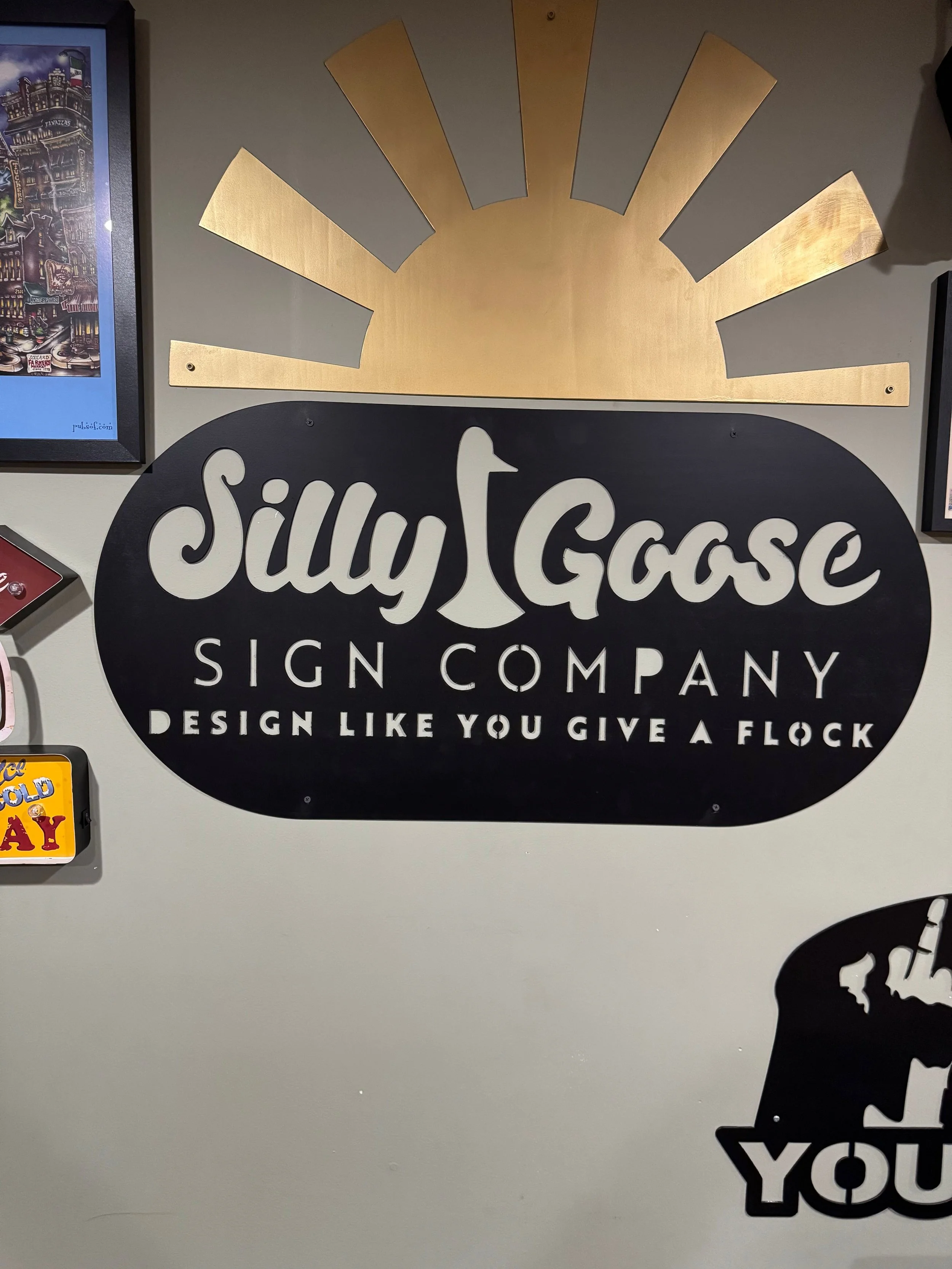 A decorative wall sign for Silly Goose Sign Company featuring a black oval background, white stylized text, and a gold sunburst design above.