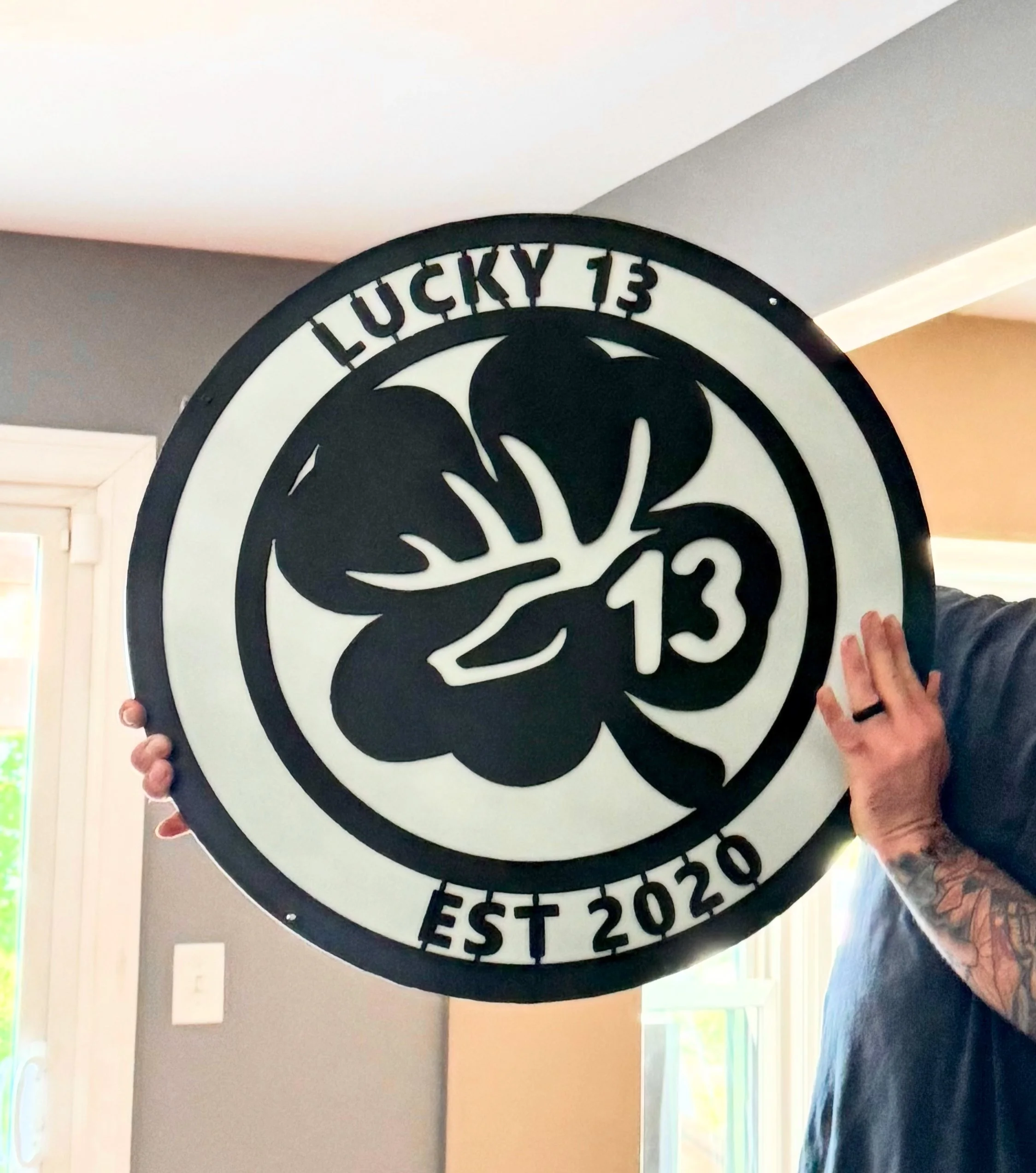 A large circular sign with a black silhouette of a Mickey Mouse head with a pumpkin face, the number 13 inside the face, and the words 'Lucky 13' at the top and 'Est 2020' at the bottom. Two hands hold the sign, one on each side.