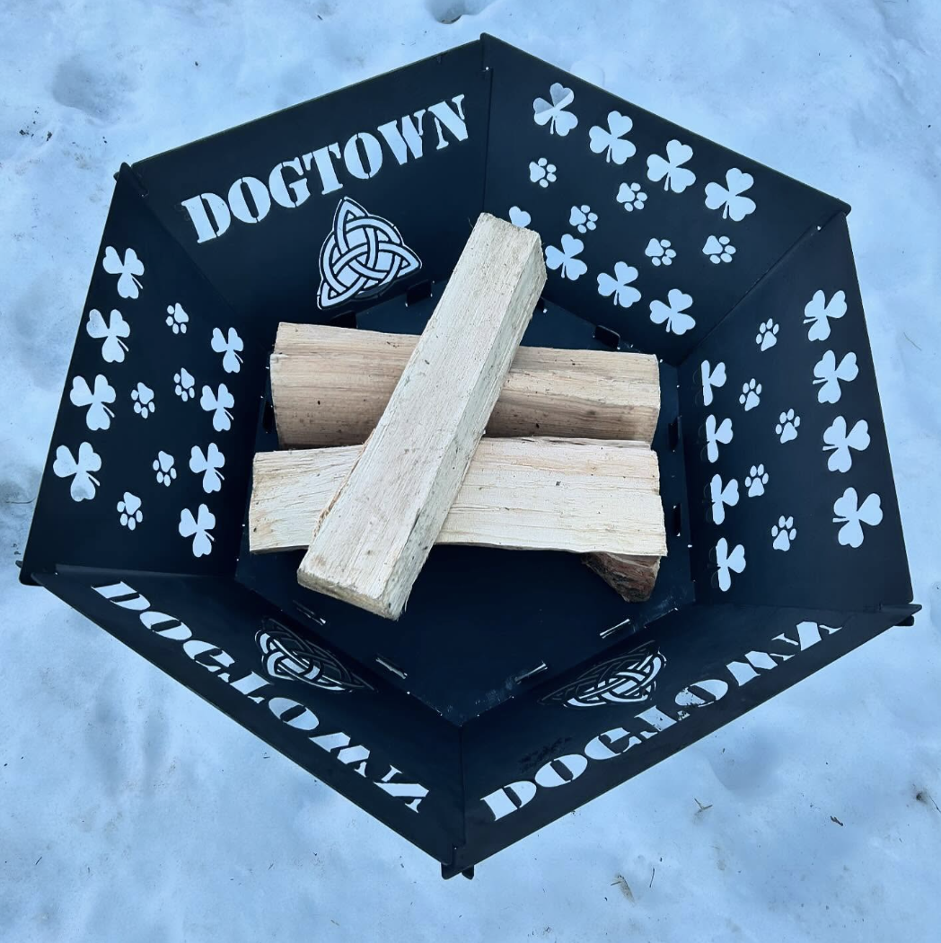 A black firepit with white shamrock and Celtic knot designs, containing several pieces of firewood and the word 'DOGTOWN' on the sides, sitting on a light-colored surface.