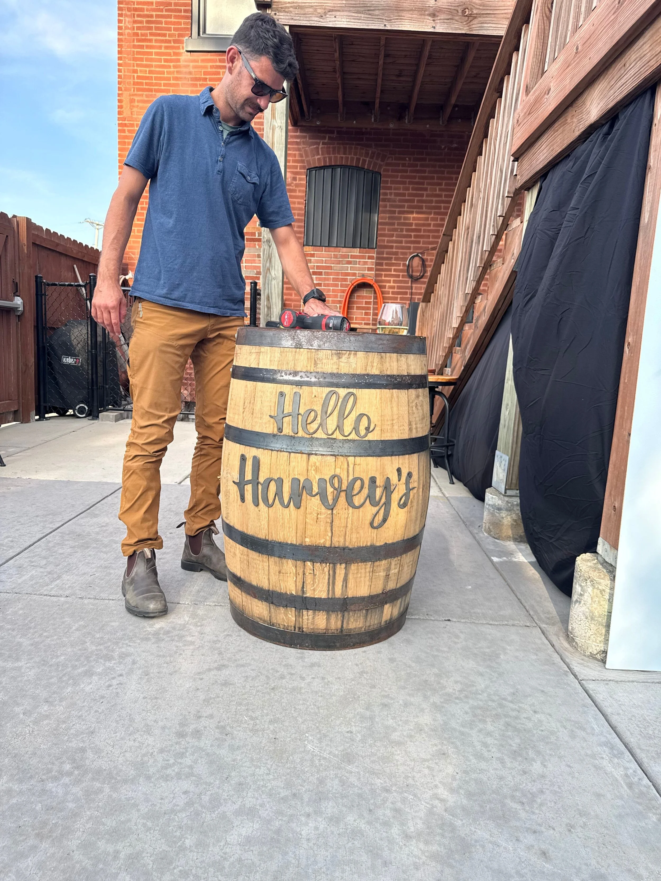 Man in sunglasses standing next to a wooden barrel with 'Hello Harvey's' written on it, outdoors in a patio area.