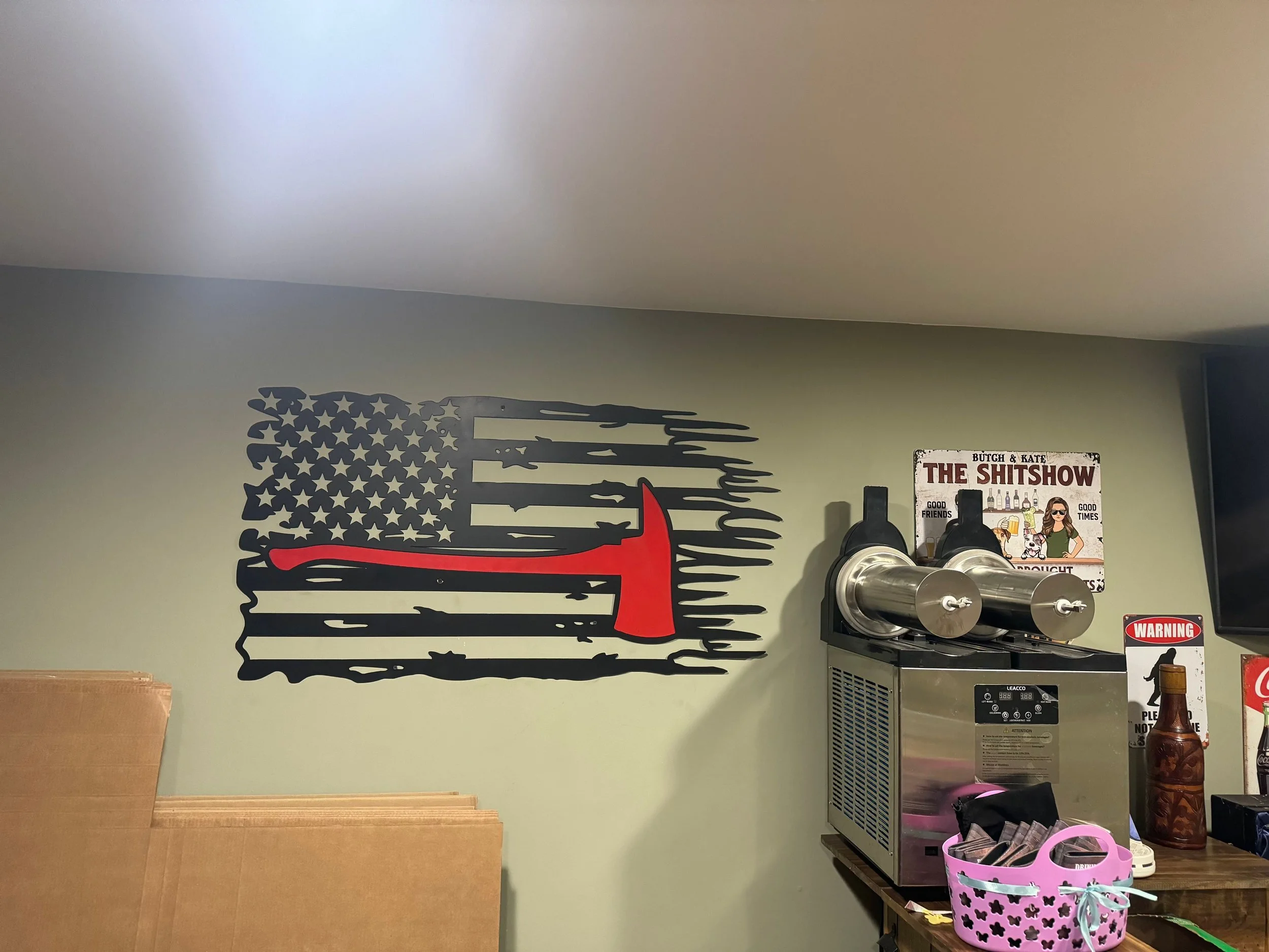 Decorative wall with a black American flag featuring a red axe in the center, a sign reading 'The Shitshow' and a drink dispenser, along with miscellaneous items on a table.