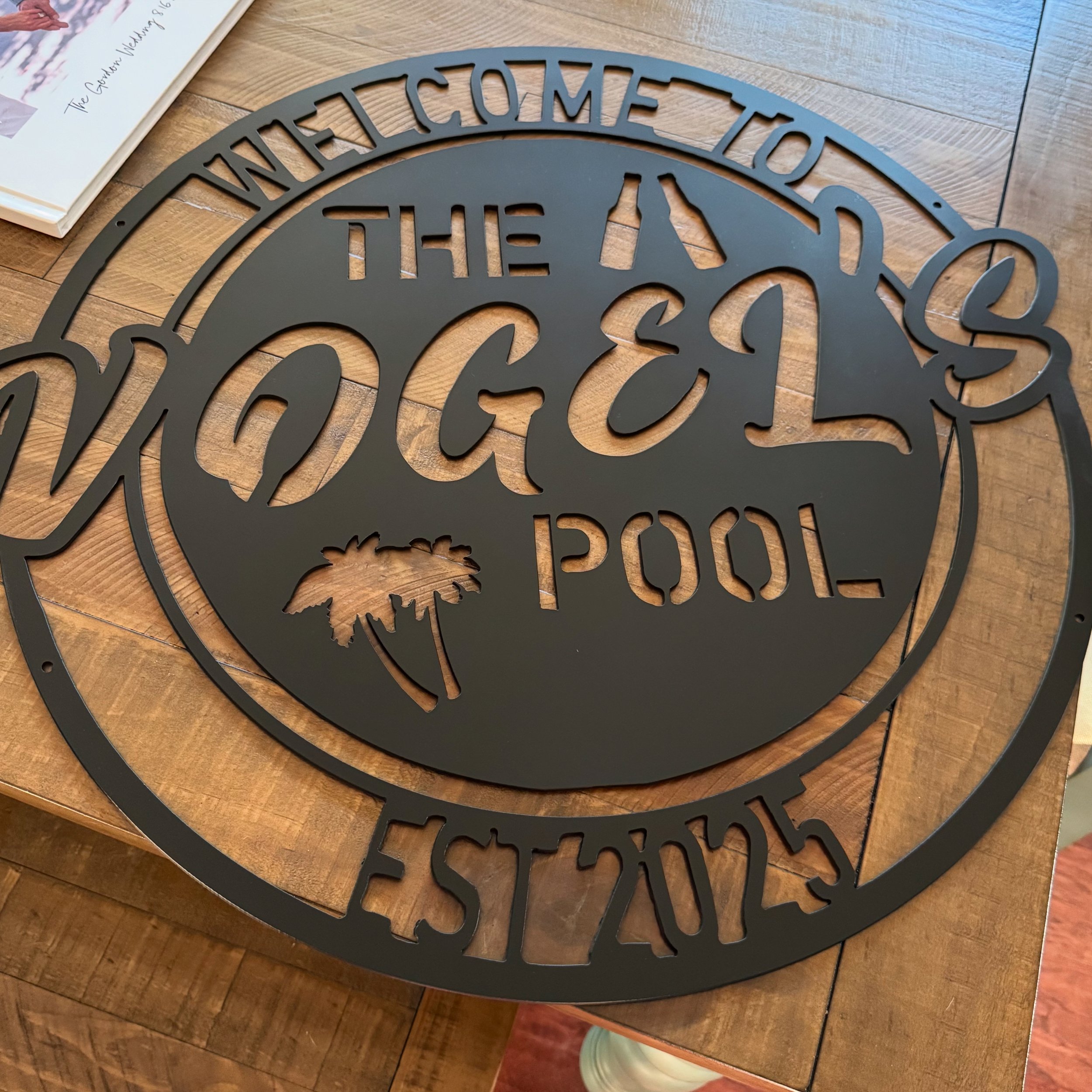 A black metal sign that says 'Welcome to the Palms Pool Festival' with a palm tree silhouette, resting on a wooden surface.