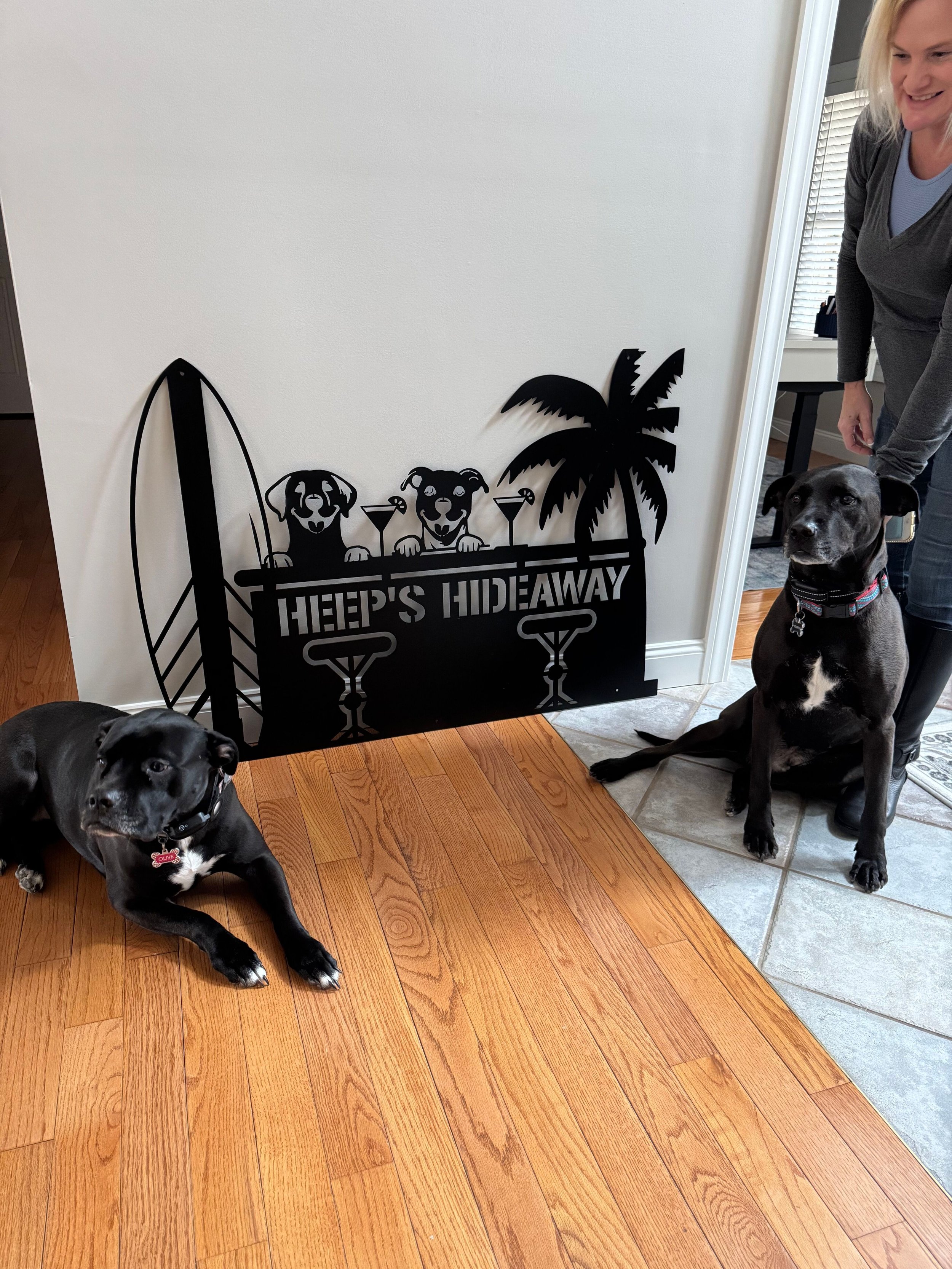 Two dogs with black fur and white markings sitting on hardwood and tile floors near a black metal sign with a surfboard design and palm tree, reading "Keep's Hideaway," with a woman standing nearby.
