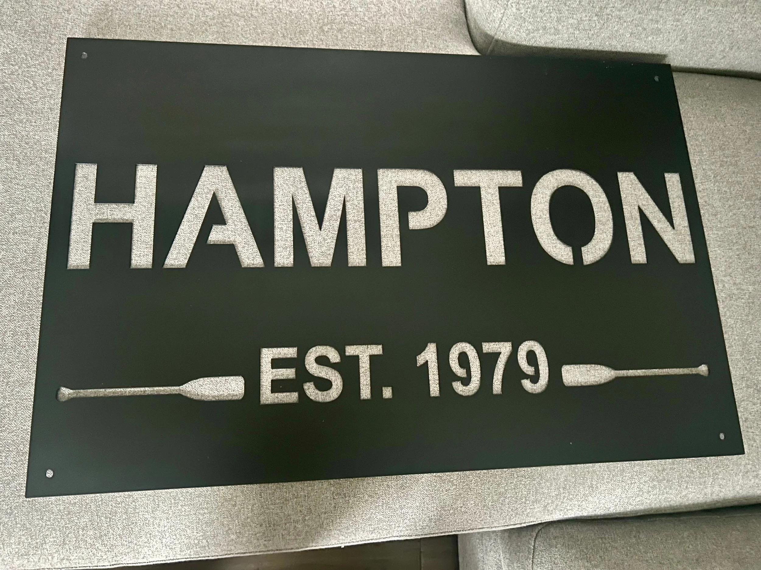 Black sign with gray text reading 'Hampton, Est. 1979', featuring two oar illustrations on either side of the text.