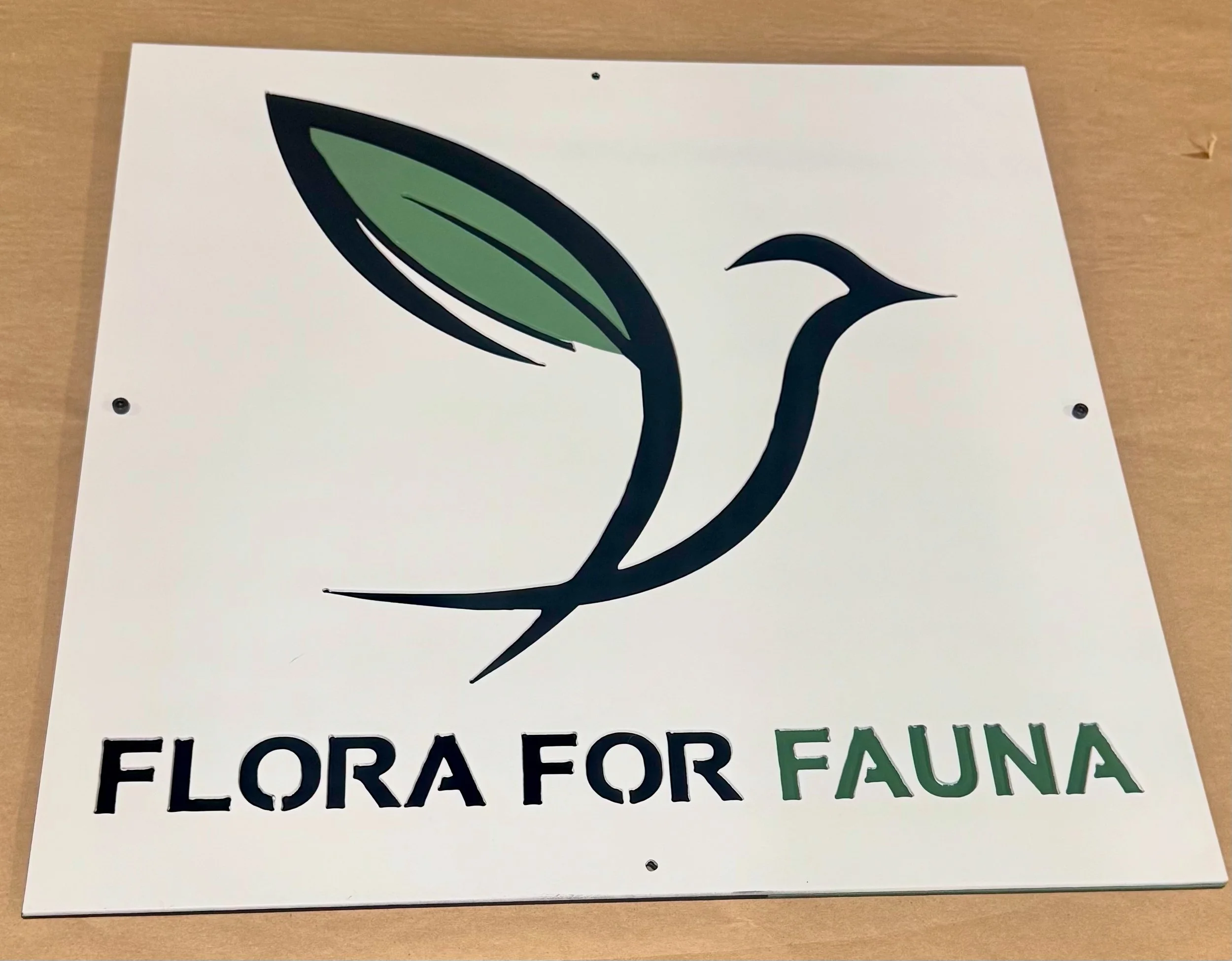 White sign with a stylized hummingbird and leaf logo, with the text 'Flora for Fauna' in black and green lettering.