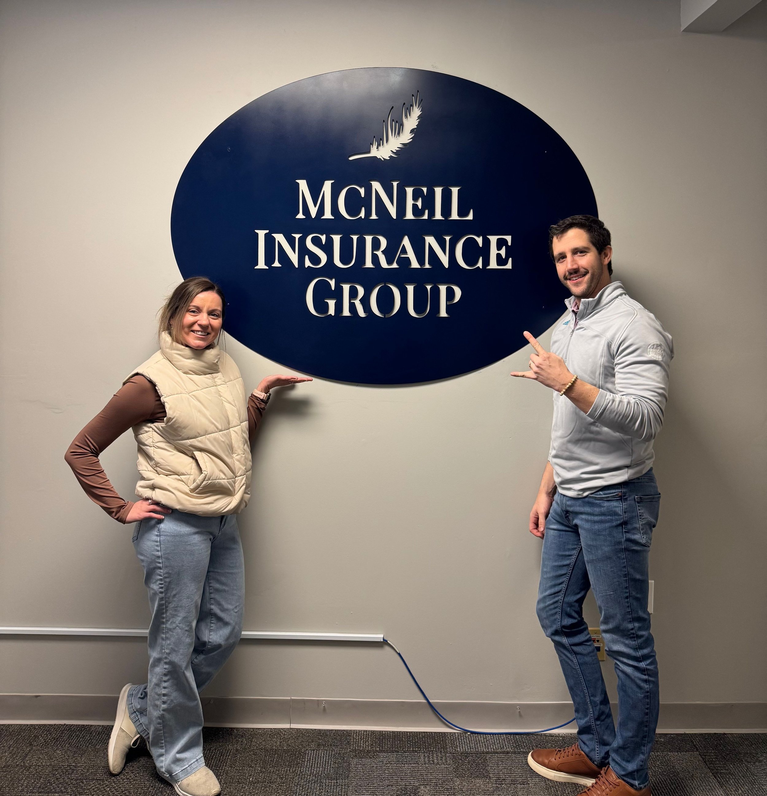 Two people standing next to a sign reading "MCNEIL INSURANCE GROUP" on a gray wall, smiling, with the woman wearing a beige vest and the man wearing a gray jacket.