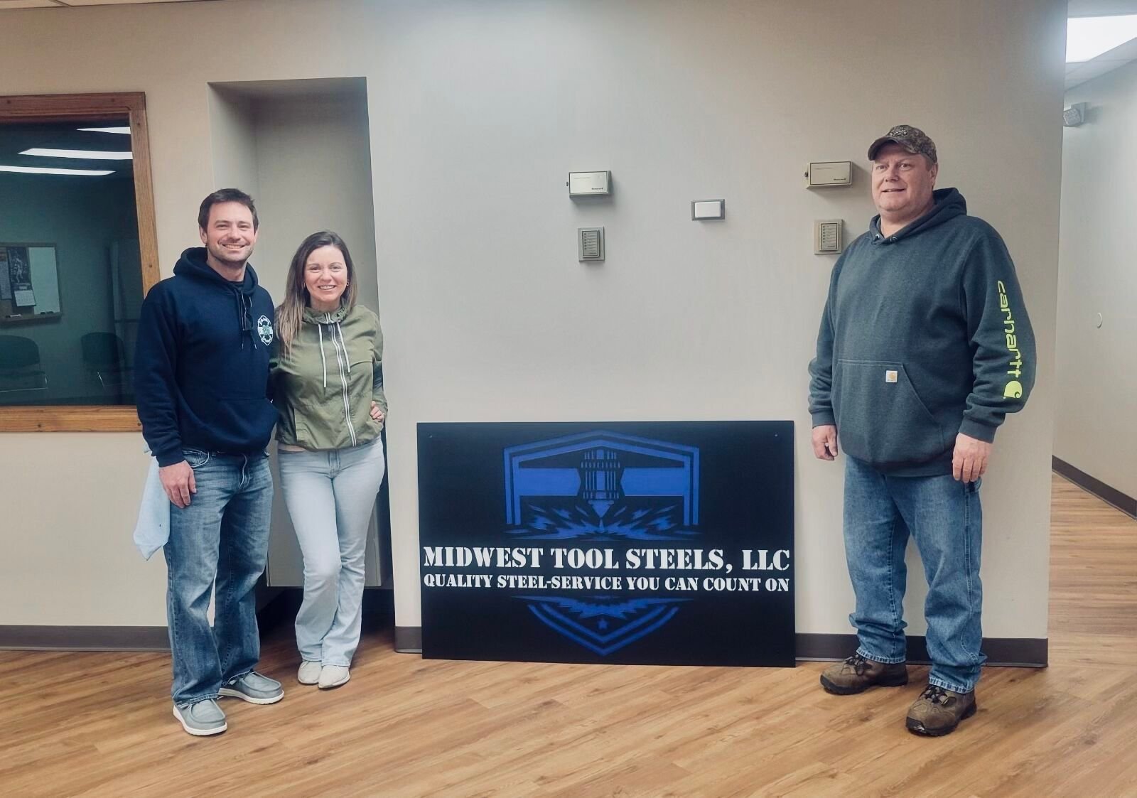 Three people standing beside a sign for Midwest Tool Steels, LLC, in an indoor setting with wood flooring and neutral walls.