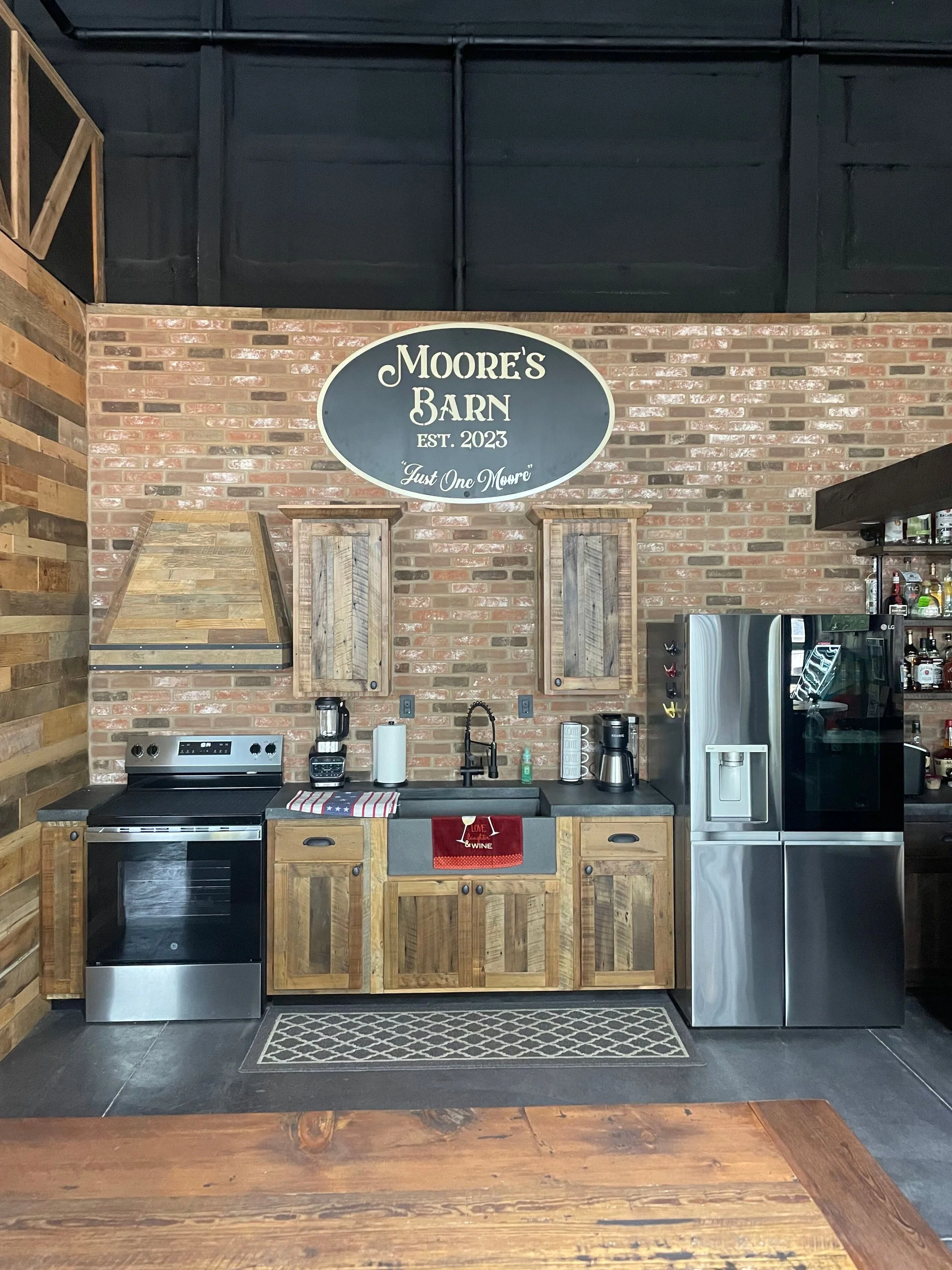 Kitchen with brick wall, wooden cabinets, stainless steel refrigerator, stove, and a sink, featuring a sign that reads 'Moore's Barn EST. 2023 Just One Moore'.