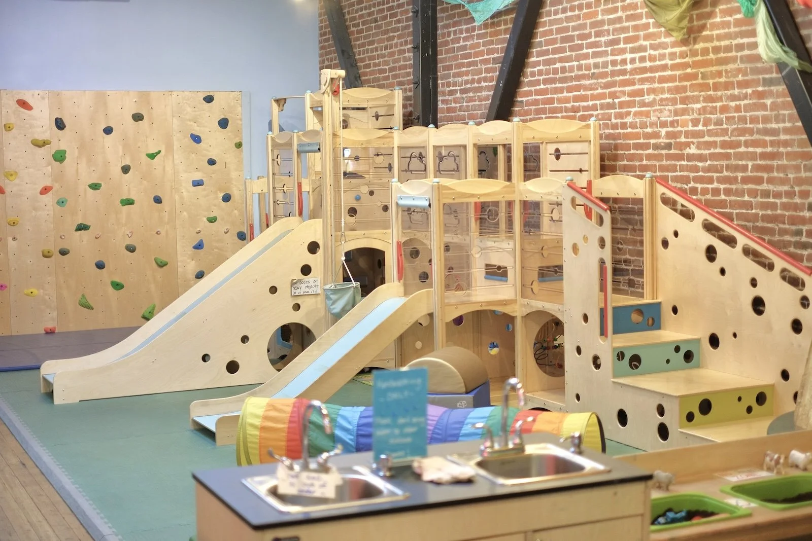 Wooden climbing structure with slide, tunnel, and kid-height play sinks in the play room