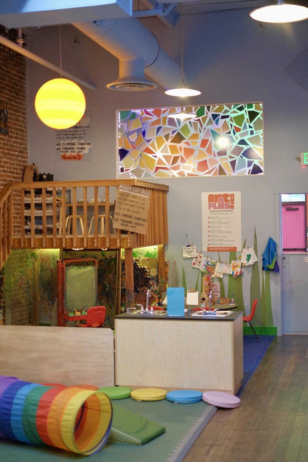 The maker loft area at Art Play Café, with a stained glass window, wooden loft, and rainbow tunnel toy