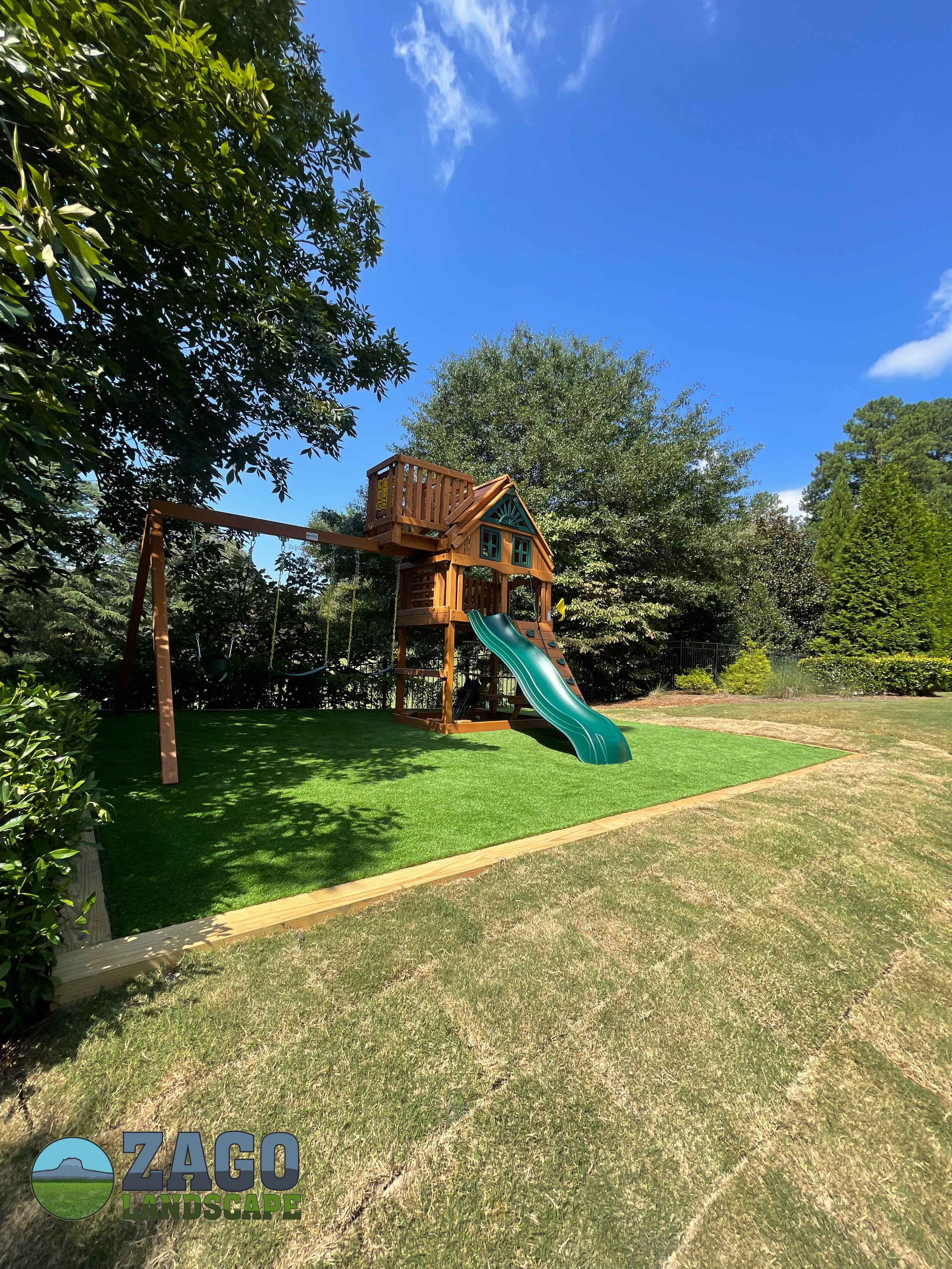 A children's playset with a green slide, a small house structure, and a wooden swing set on artificial turf, surrounded by trees and bushes under a partly cloudy blue sky.