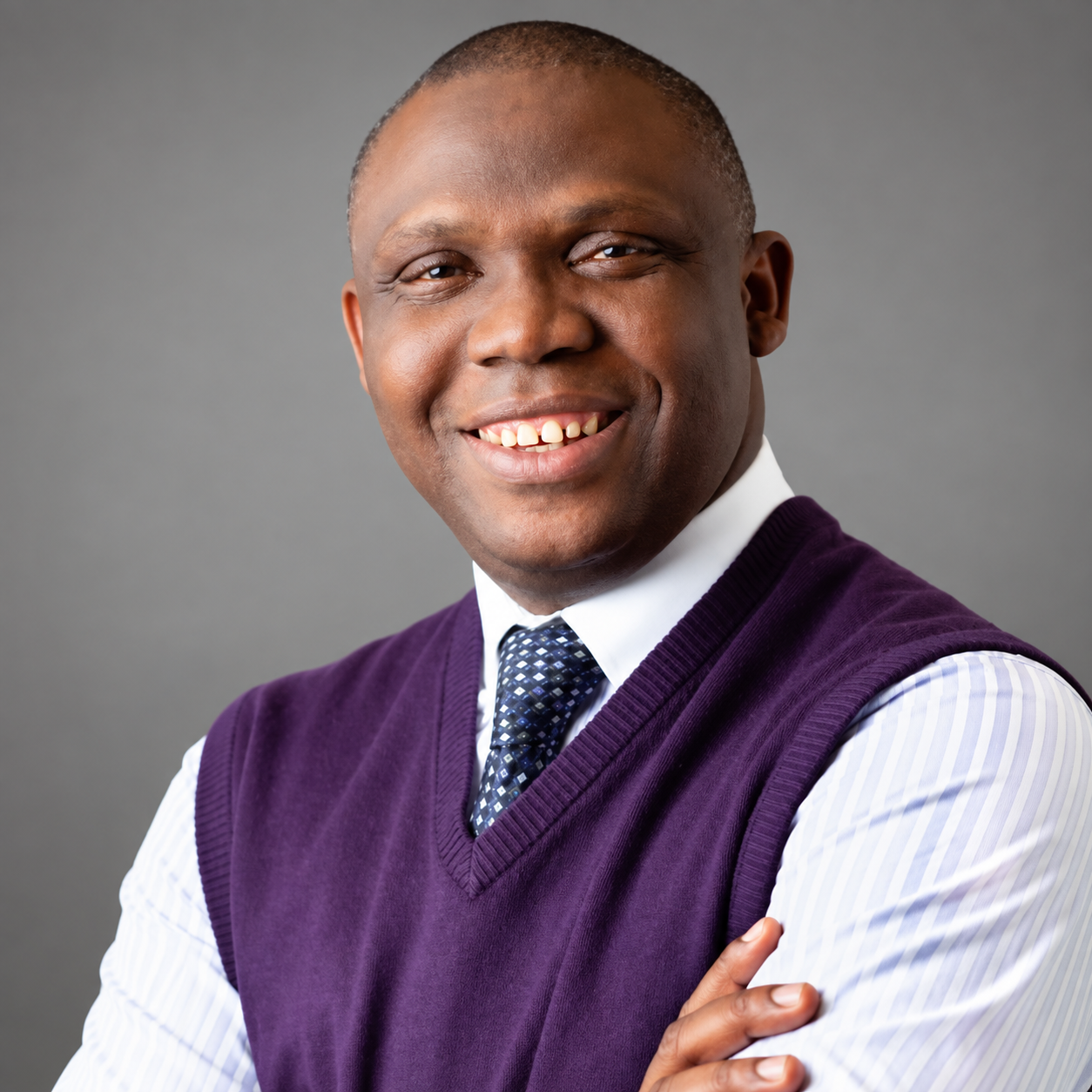 Portrait of a smiling African American man with short hair, wearing a white shirt, purple vest, and patterned tie, against a gray background.
