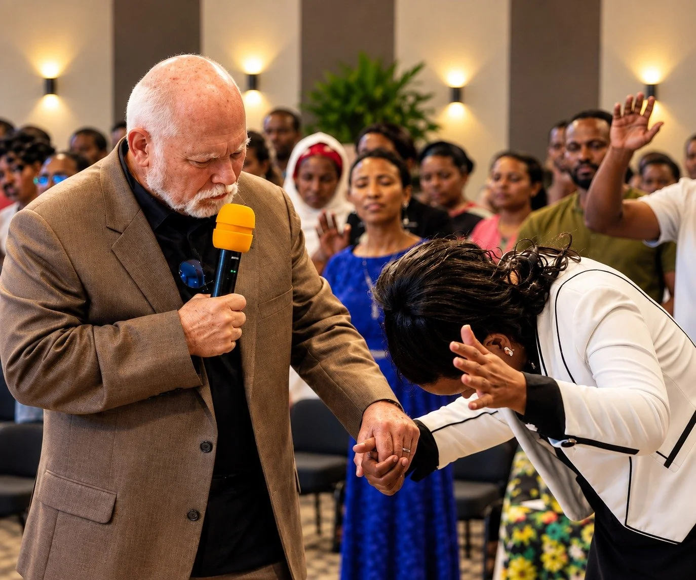 A religious ceremony where a man in a brown blazer is holding a microphone and praying over a woman with dark hair, who is bowing her head and holding his hand. Several people are gathered in the background, watching the ceremony.