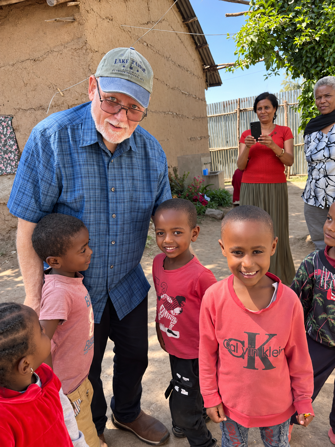 An elderly man with glasses and a cap is holding hands with young children outdoors. The background features a dirt ground, a mud house, a fence, and some women watching and taking photos.