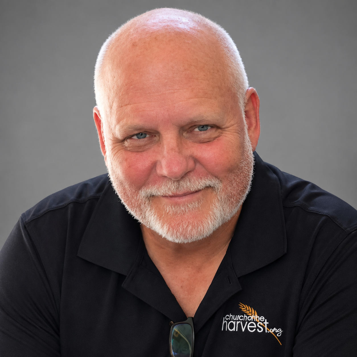 A smiling older man with a beard and blue eyes, wearing a black shirt with 'church on the harvest.org' logo, against a gray background.