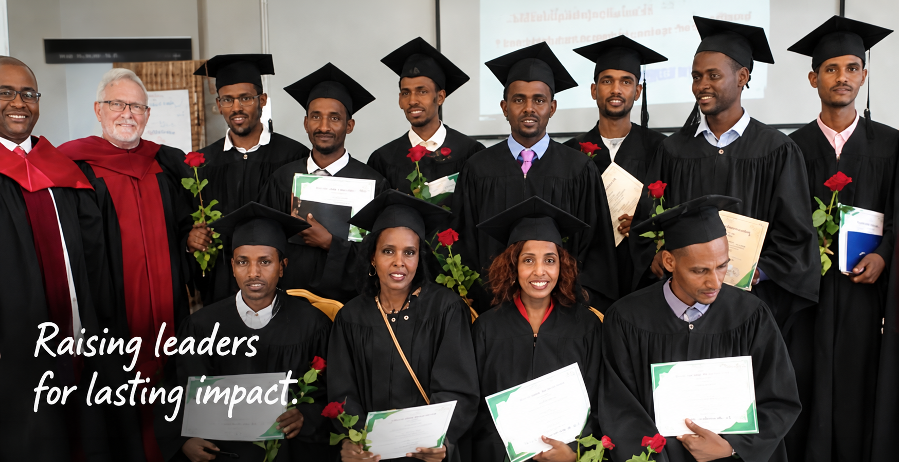 A group of graduates wearing caps and gowns, holding diplomas and red roses, posing with faculty members, at a graduation ceremony.