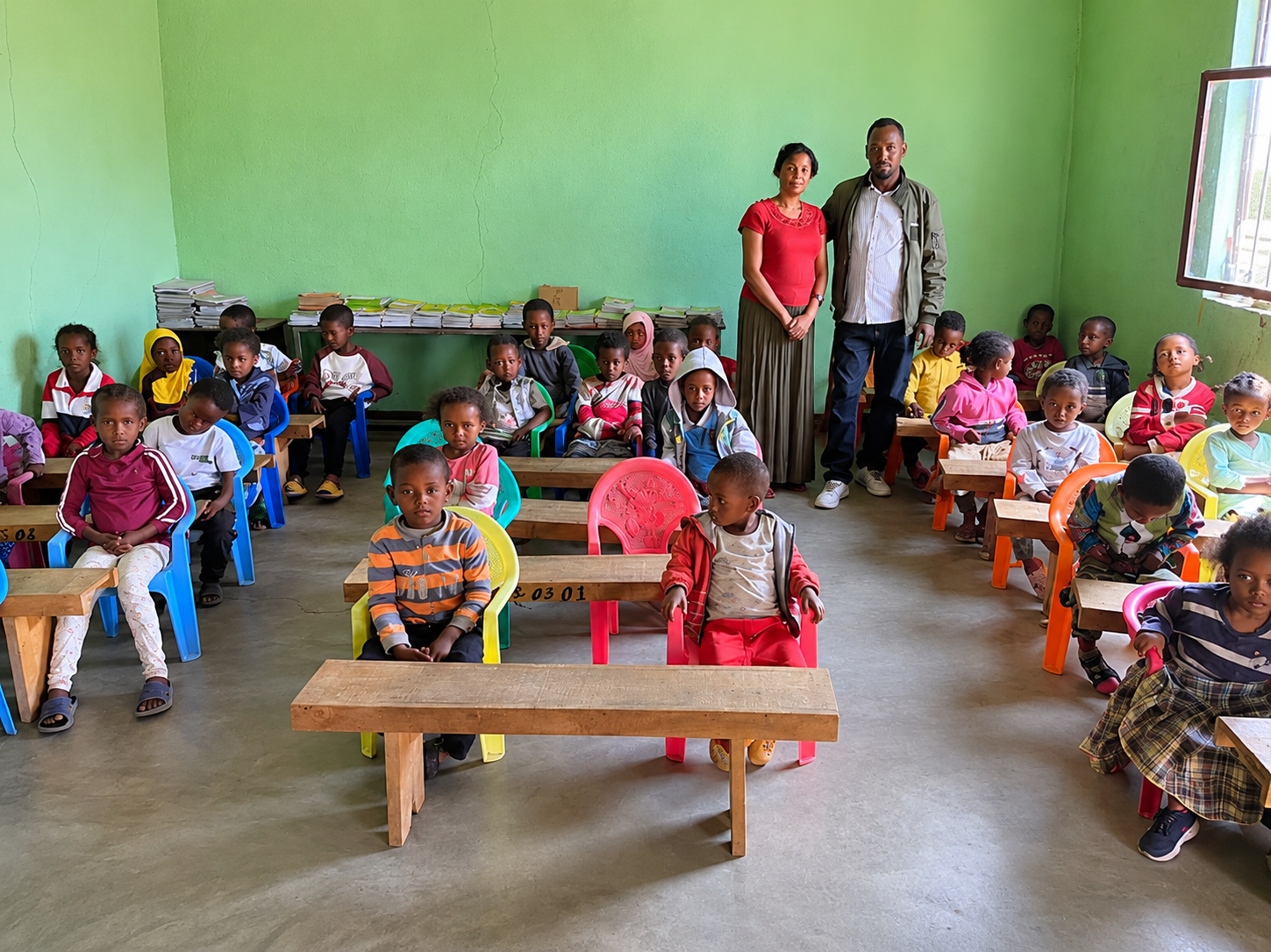 Children sitting in a classroom with two teachers standing at the back, green painted walls, and a window on the right side, with colorful plastic chairs and desks.