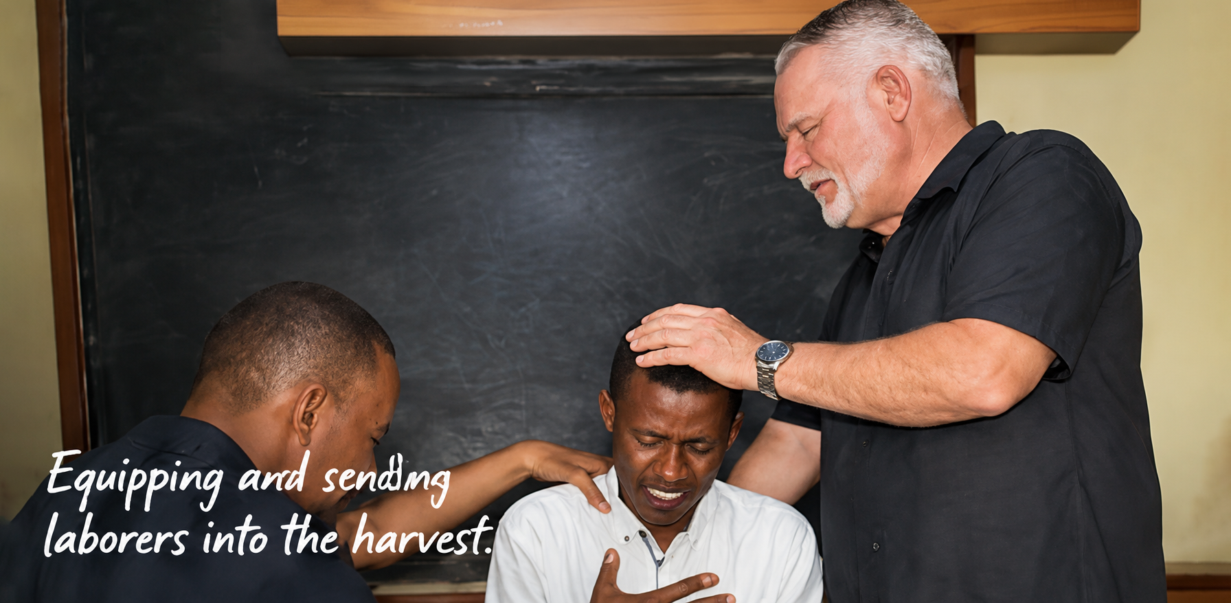 A man with grey hair and a black shirt is laying his hand on the head of a young man with a distressed expression, while another young man prays with his hand on the shoulder of the distressed man, in front of a blackboard. The text overlay says, 'Equipping and sending laborers into the harvest.'