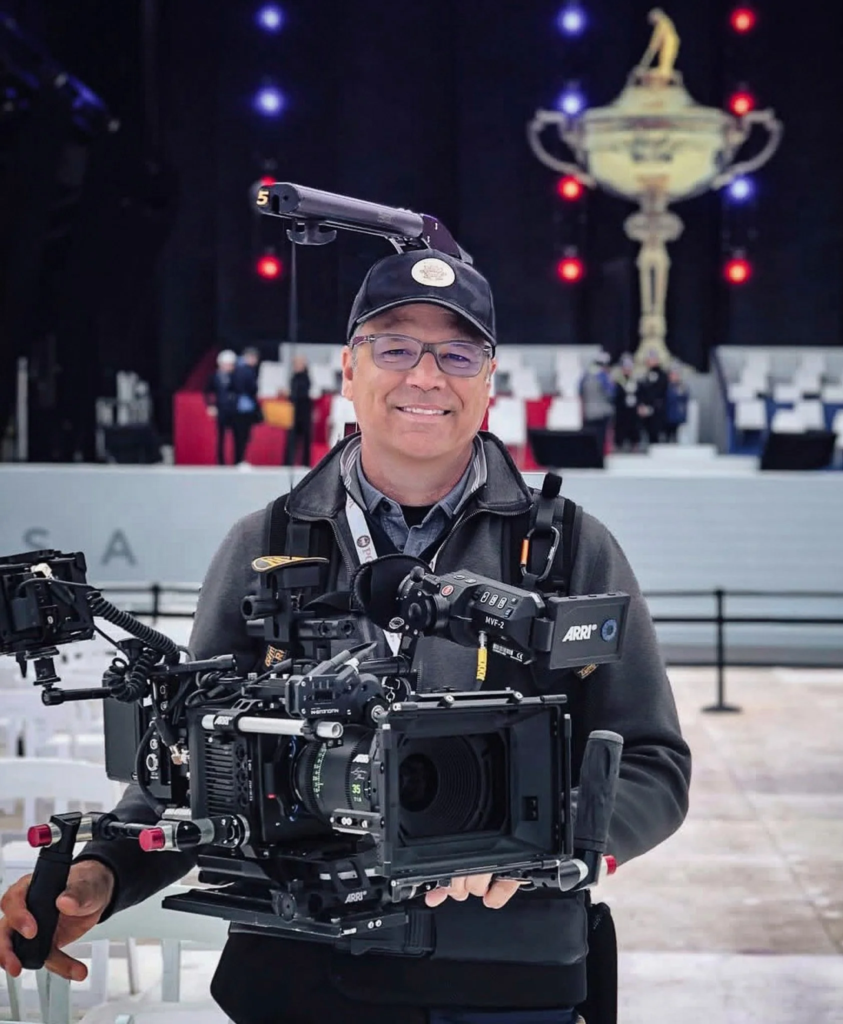 A man in glasses and a black cap holding a professional camera rig in an indoor venue with a large trophy sculpture and people on stage in the background.