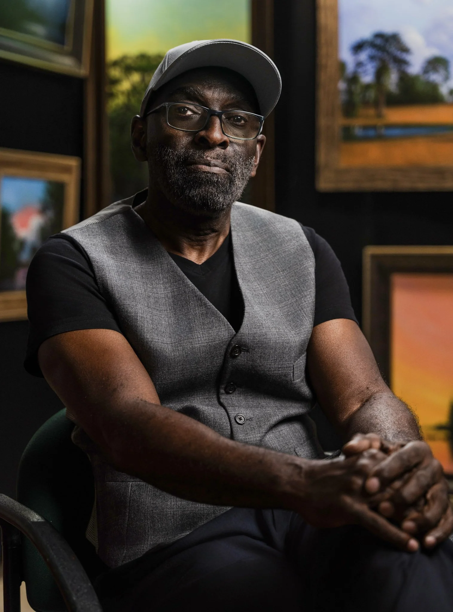 A man with glasses, a gray cap, and a short beard, wearing a gray vest over a black shirt, sitting indoors with his hands clasped, in front of framed paintings.