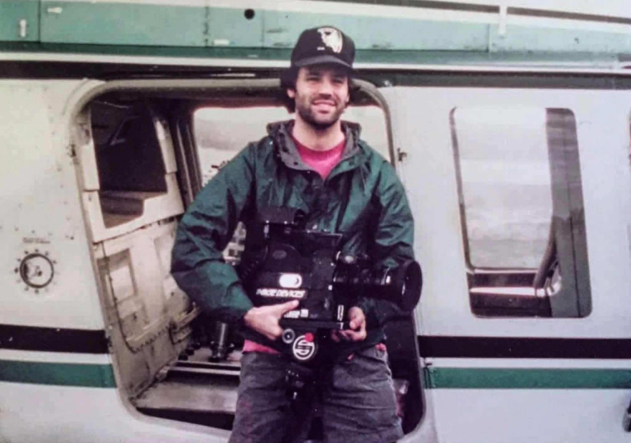 A man standing in the open door of a helicopter, holding a large camera, wearing a baseball cap, green jacket, and pink shirt underneath.