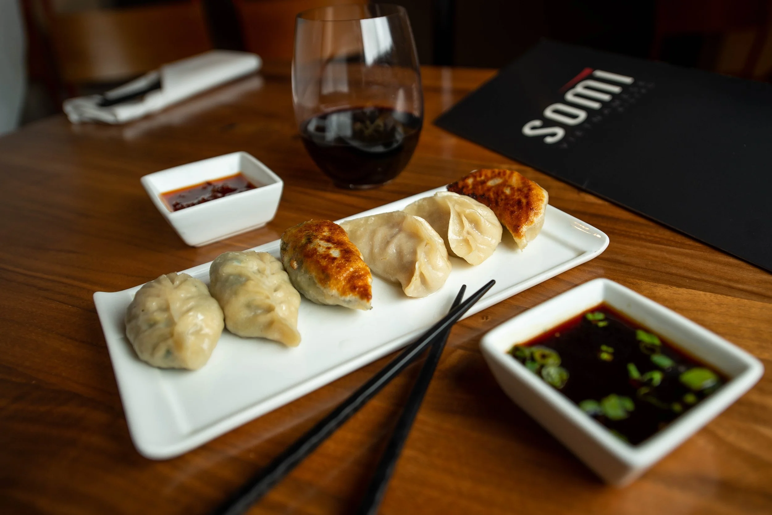 Dumplings with dipping sauces on a white plate, chopsticks, and a glass of red wine on a wooden table.
