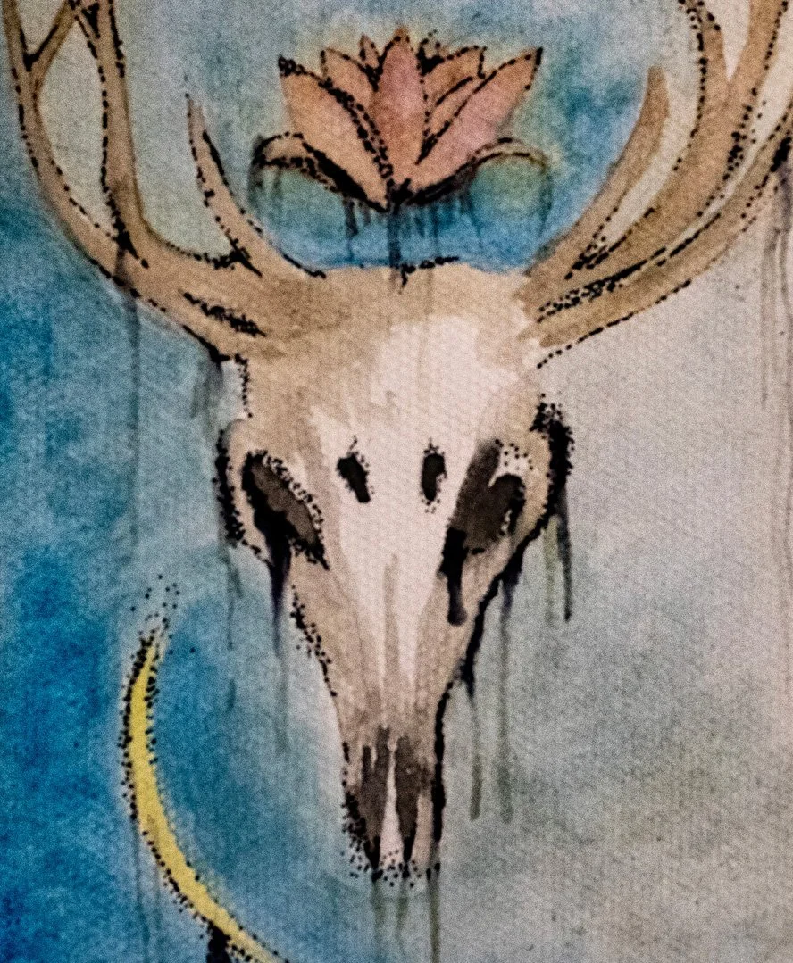 Lotus and Bone. A watercolor painting of a deer skull featuring prominent antlers, set against a textured blue background evoking the night sky. A soft pink lotus flower blooms from the center of the skull's forehead, symbolizing love and spiritual c