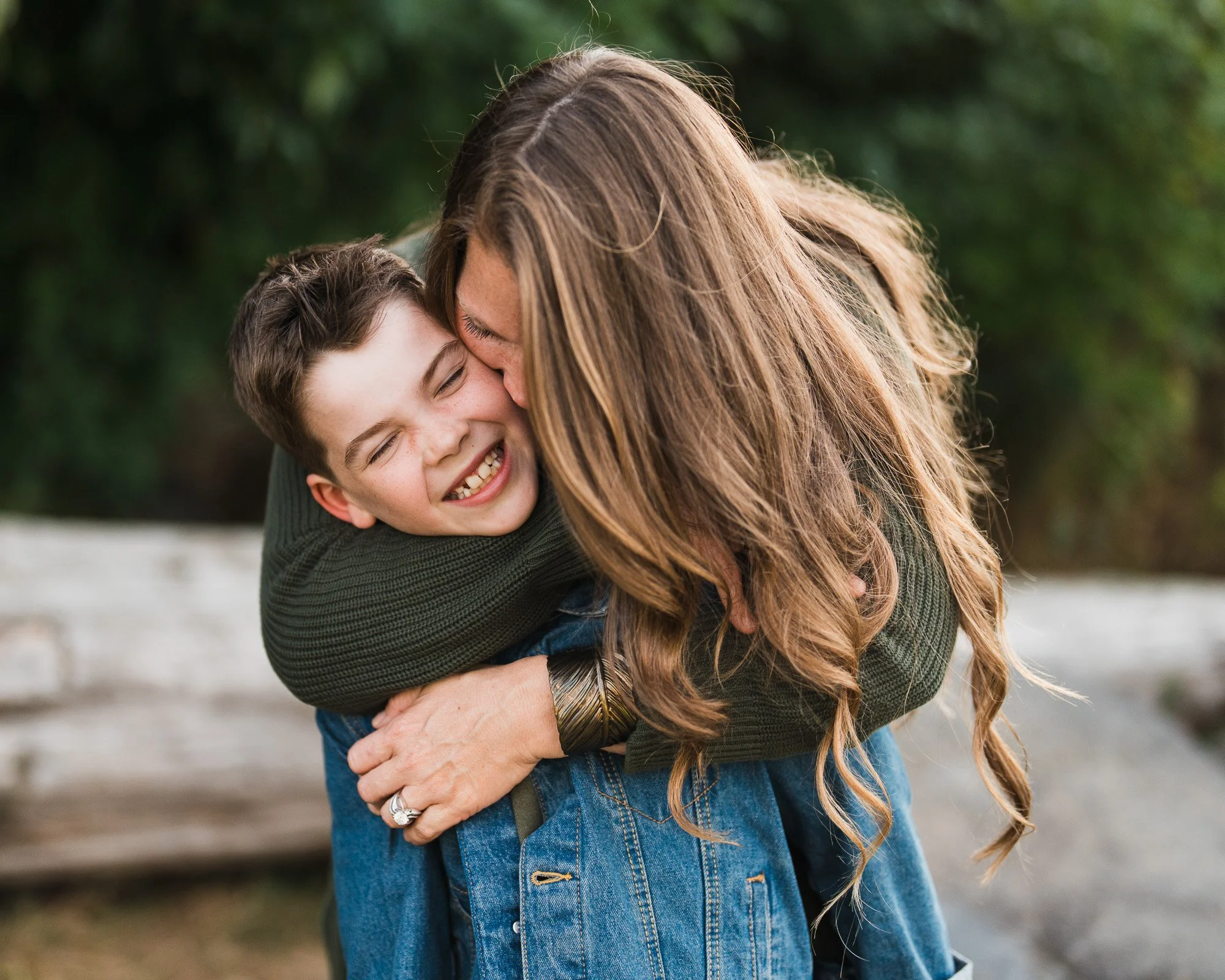 Mother kisses her smiling child’s cheek while wrapping him in a hug at a beach during a family photoshoot at Sammamish Lake.