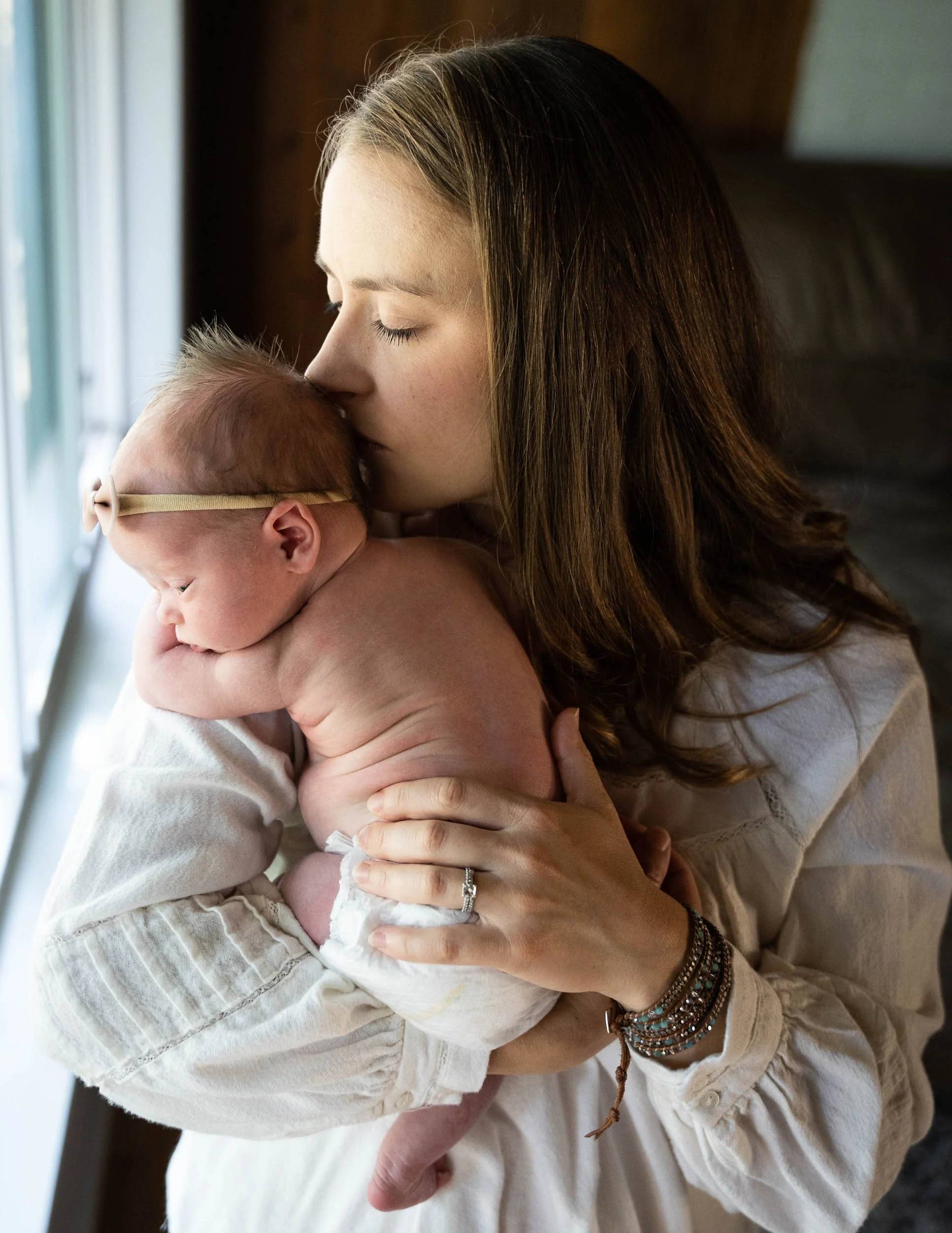 A new mom is gently kissing her baby's head near the window during a newborn photography session in Kirkland WA. 