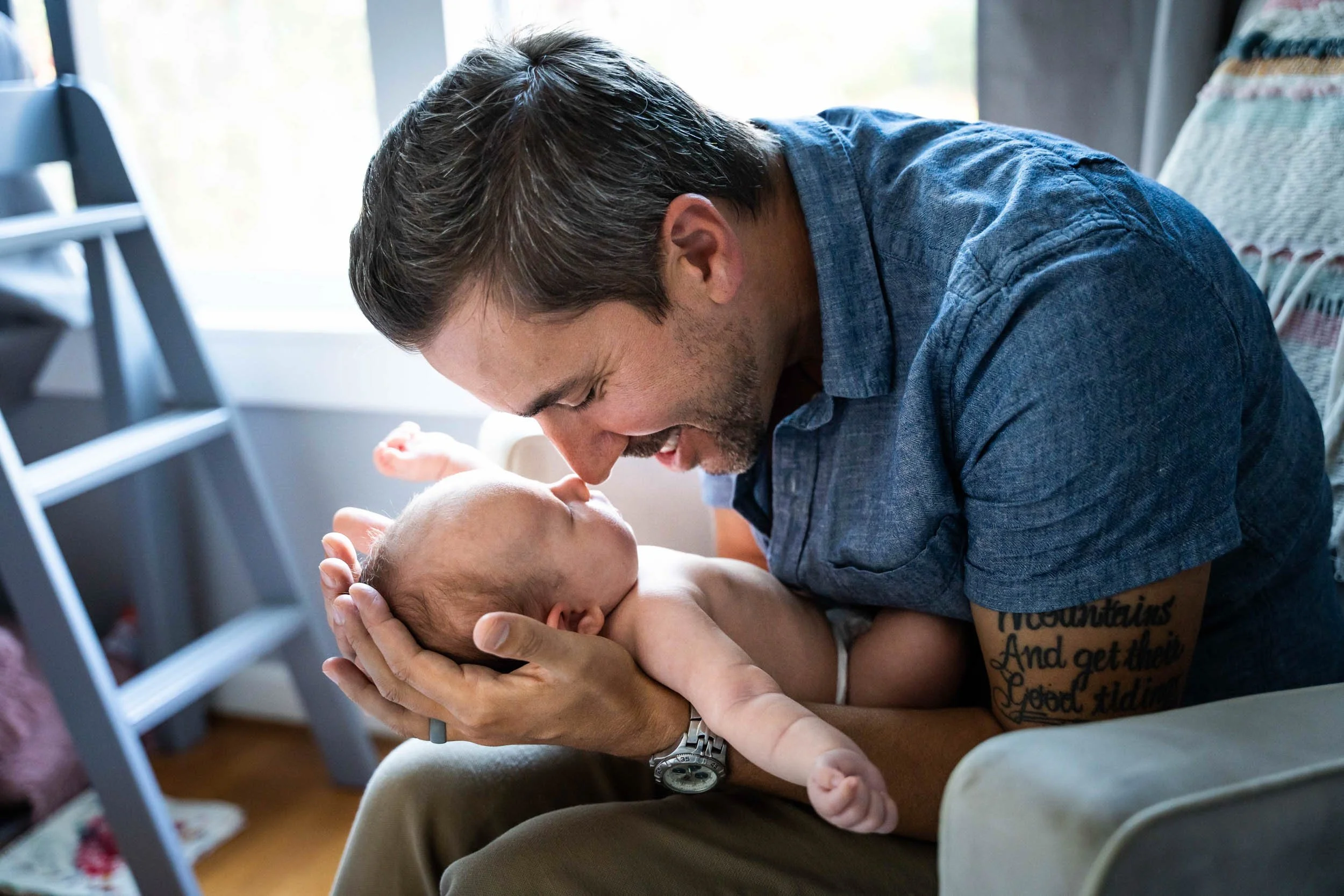 Father cradles his newborn in both hands and touches noses with the baby beside a window at a newborn photo shoot in Bellevue, WA.
