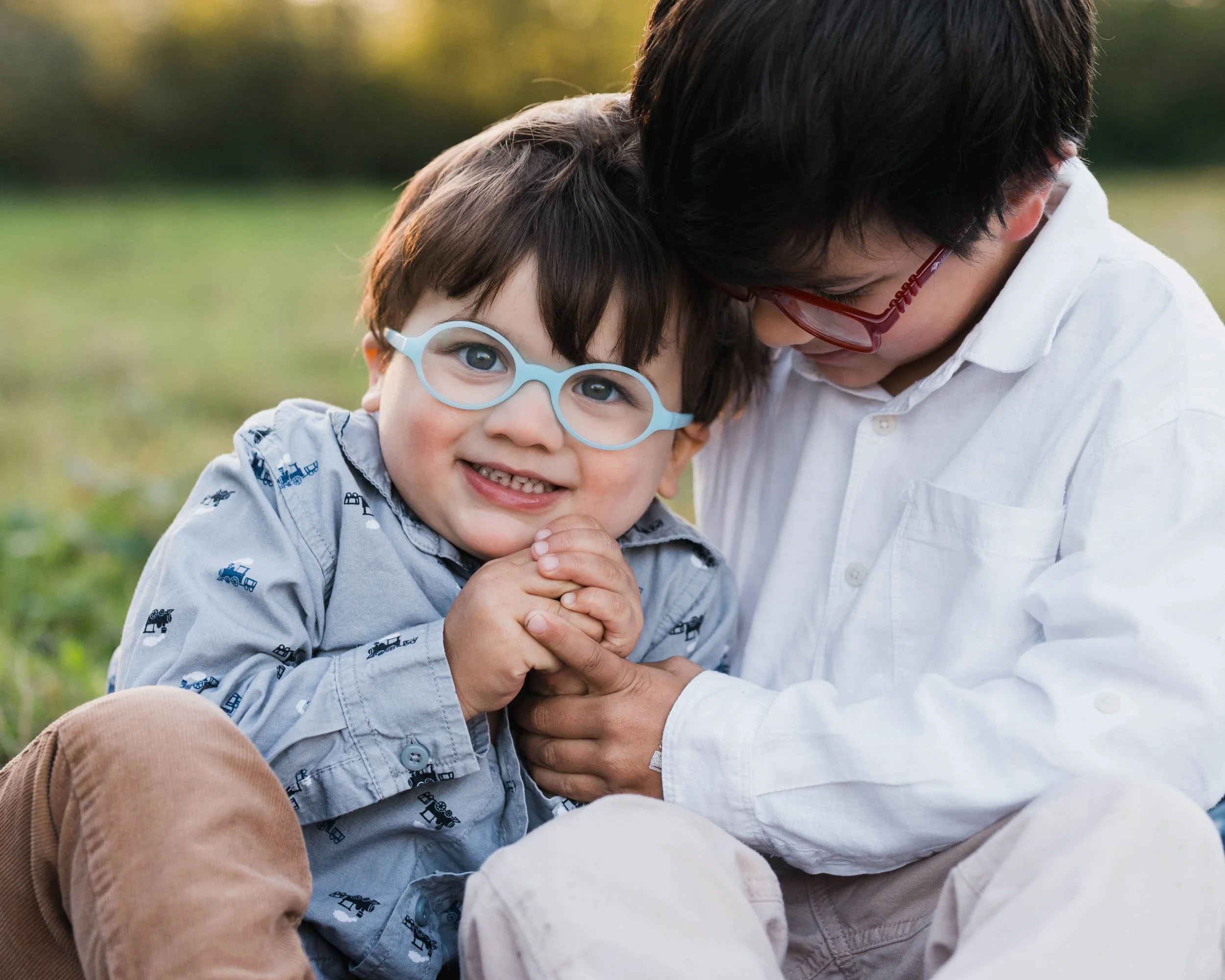 Smiling child in light blue glasses clasps hands with older brother who leans close outdoors in a field infront of Mt Si in North Bend.