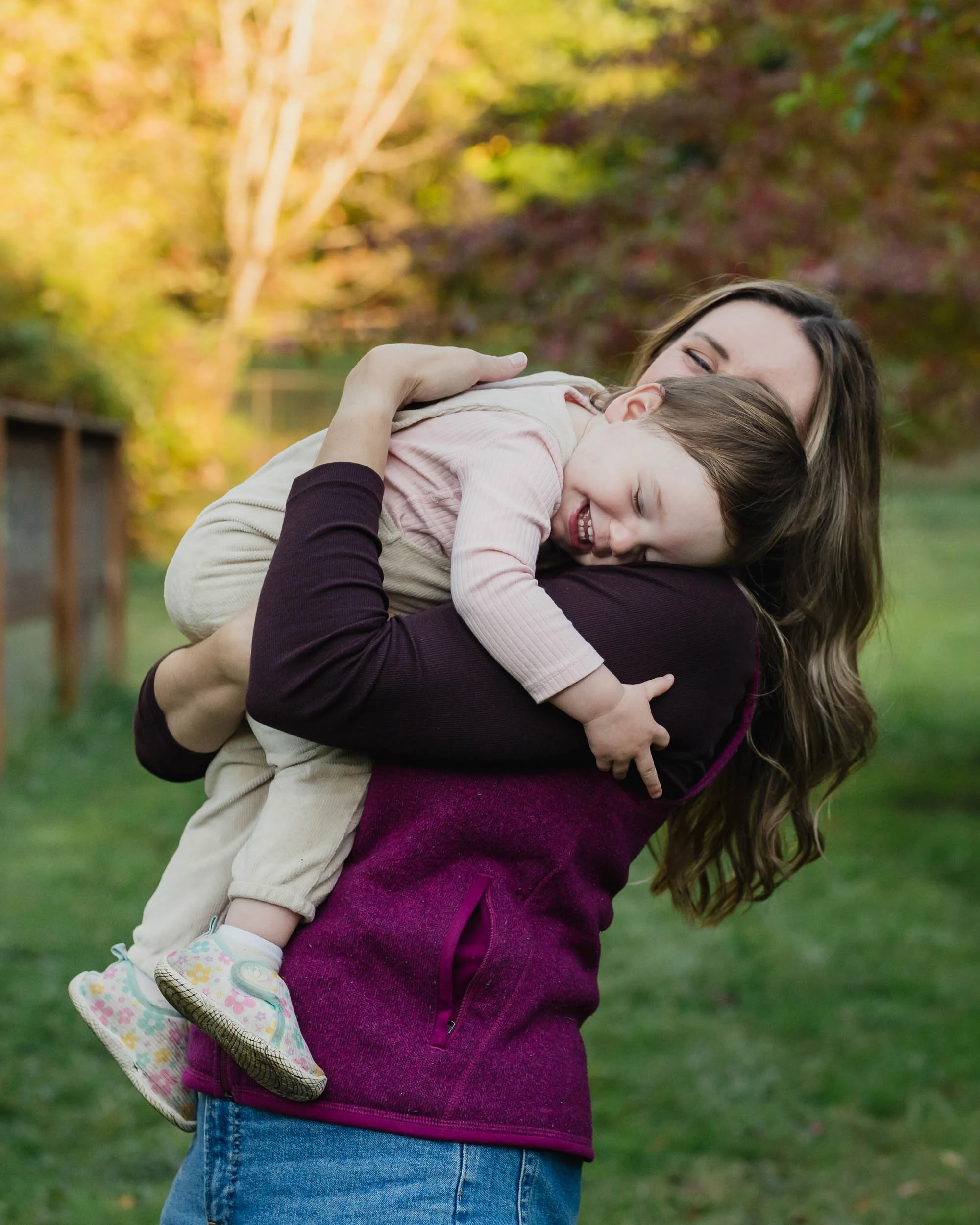 Mother holds her laughing child over her shoulder among autumn trees in Redmond family photo session.