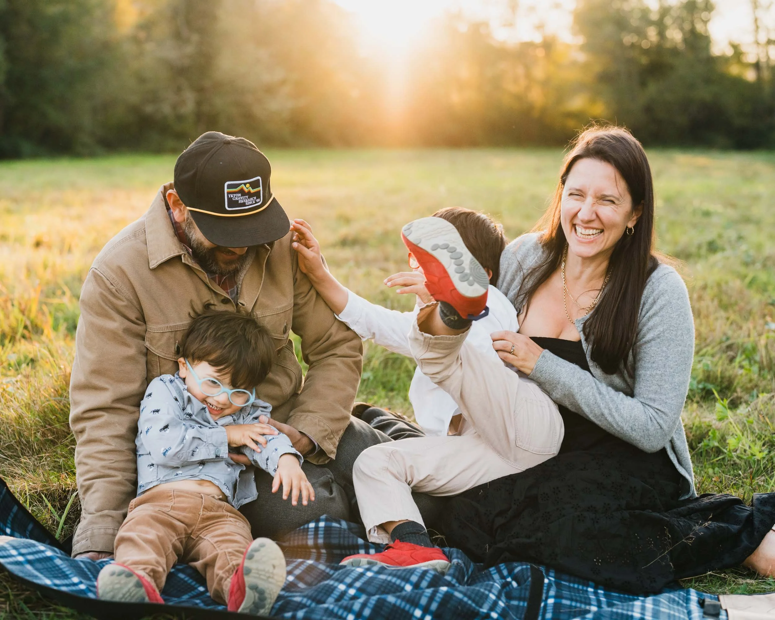 Family of four laughs together on the grass at golden hour as one child kicks a foot into the air.