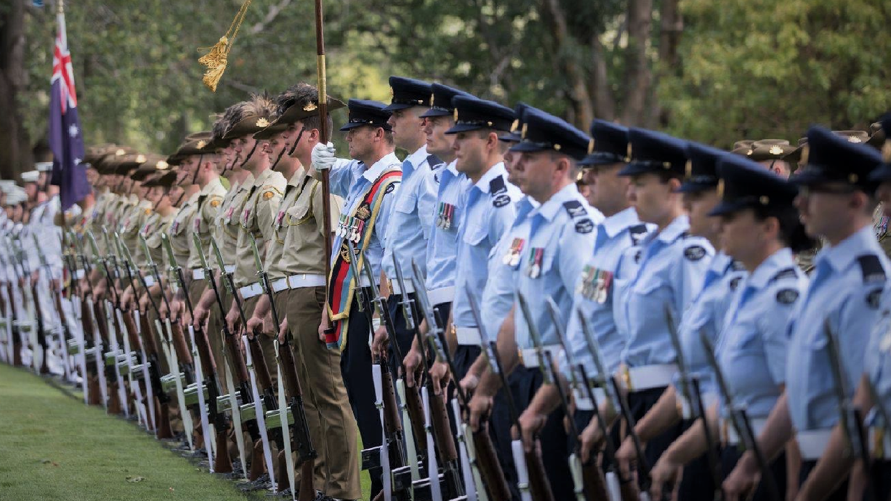 Line of uniformed military personnel standing at attention during a ceremony outdoors, with trees in the background. Department of Veterans Affairs (DVA)