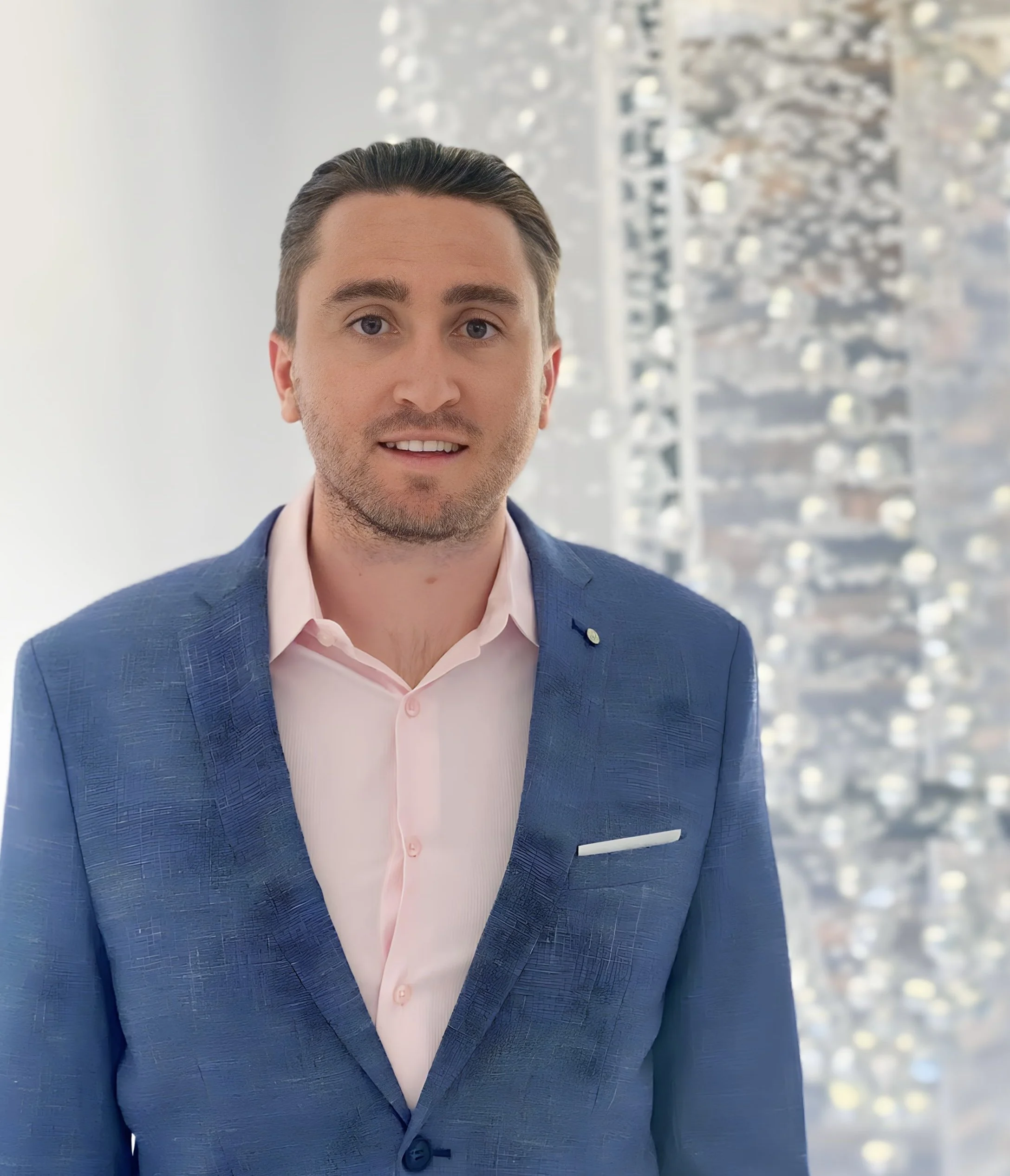 A man with dark hair and wearing a blue suit with a pink dress shirt, standing indoors with a cityscape visible through a window behind him. Department of Veterans Affairs (DVA)