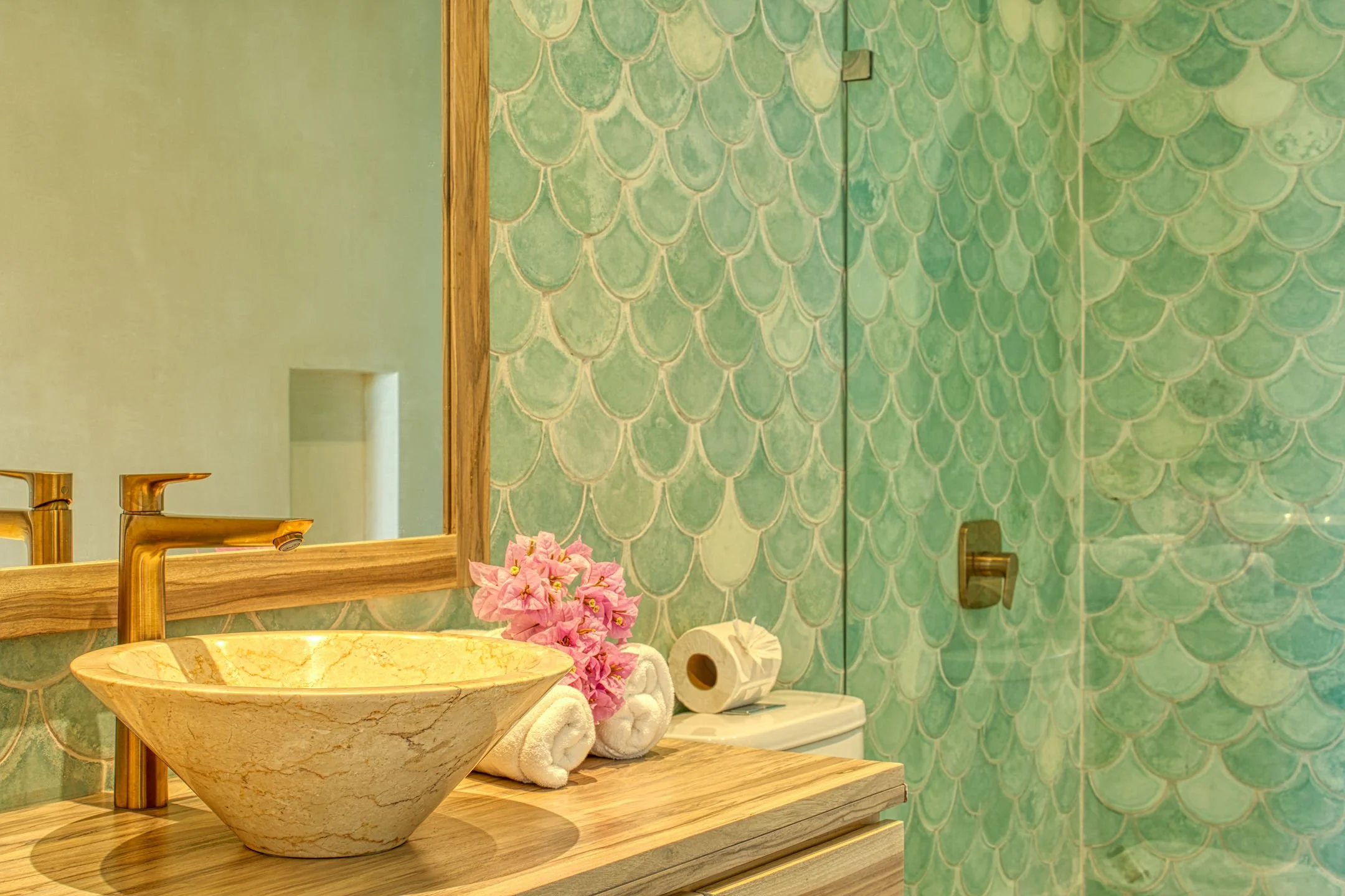 Bathroom with a marble sink, pink flowers, and towels, with a green scalloped tile wall and a mirror.