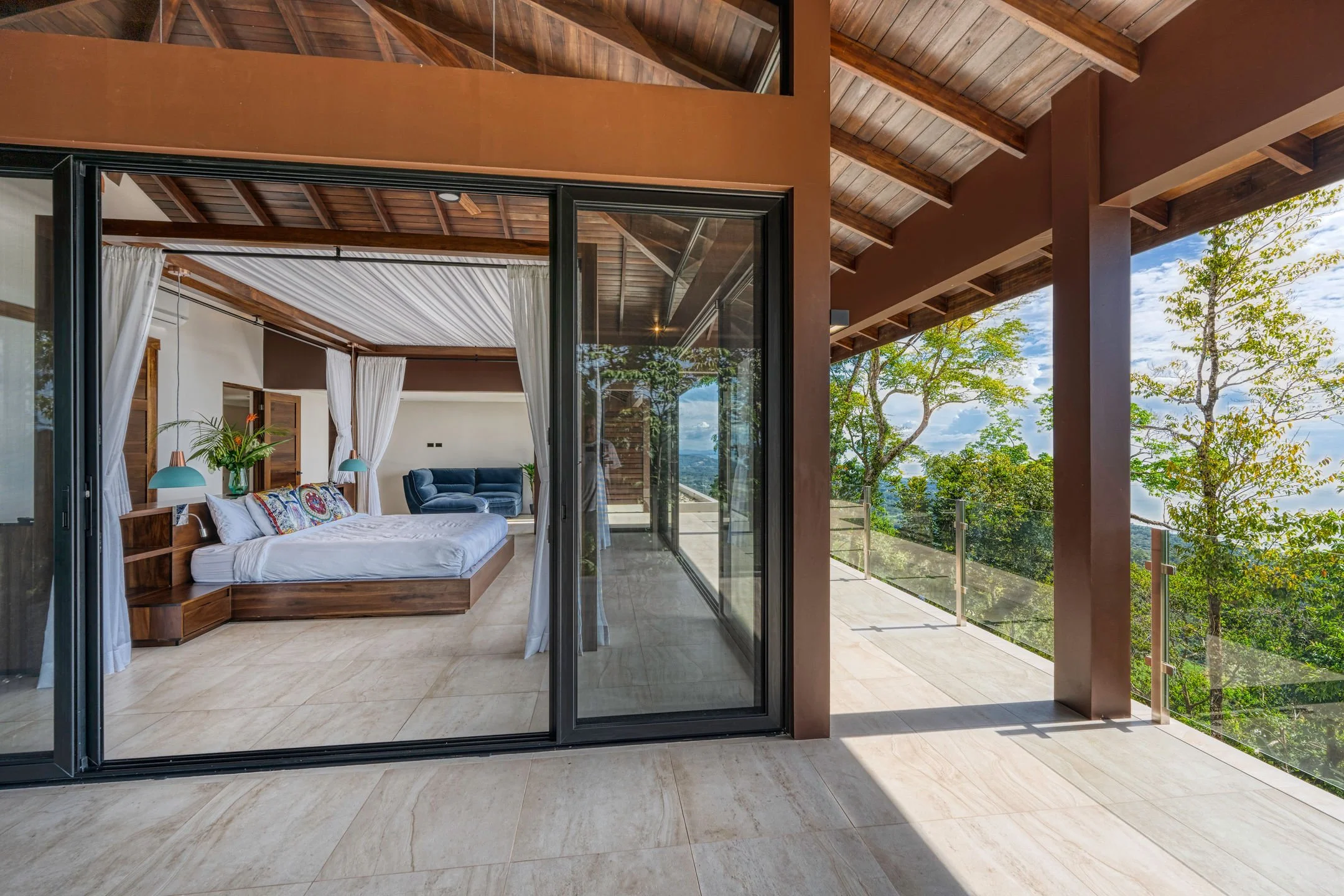 Modern bedroom with sliding glass door open to a balcony surrounded by trees and a blue sky.