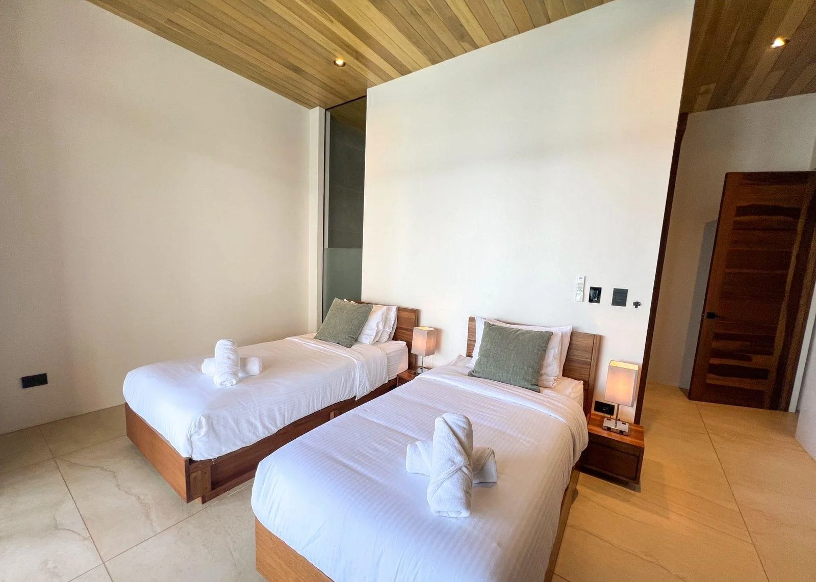 A hotel room with two twin beds decorated with white linens and green pillows, featuring wood headboards. There are bedside tables with lamps, and towels folded on each bed. The room has a wooden ceiling, cream-colored walls, and tiled floors. A wooden door is visible on the right side.