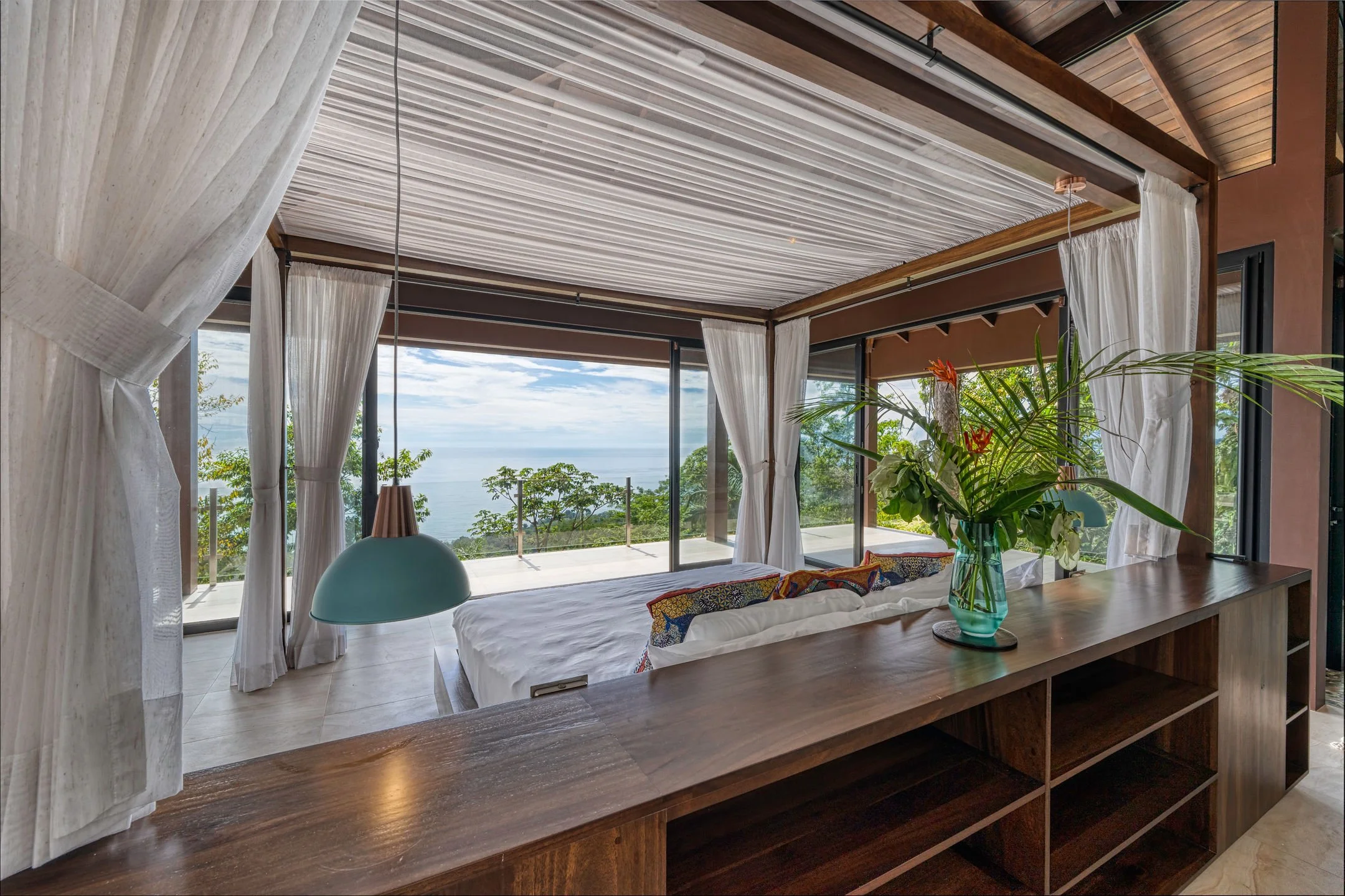 A bedroom with a bed facing large windows overlooking the ocean and trees, decorated with a bouquet of flowers on a wooden console in the foreground, draped curtains, and a hanging pendant light.