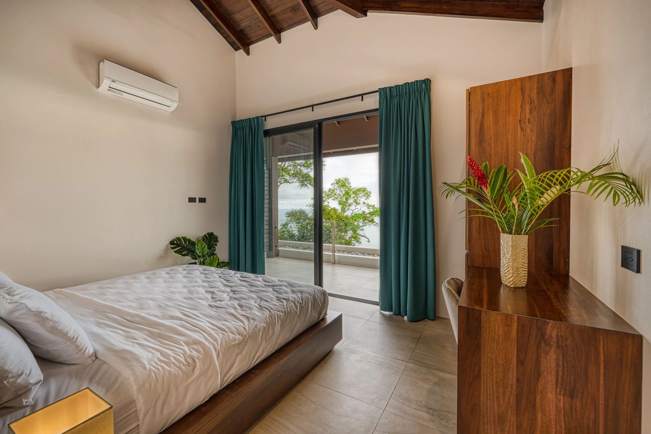 Bedroom with large bed, sliding glass door to balcony with ocean view, teal curtains, wooden ceiling, and potted plant.