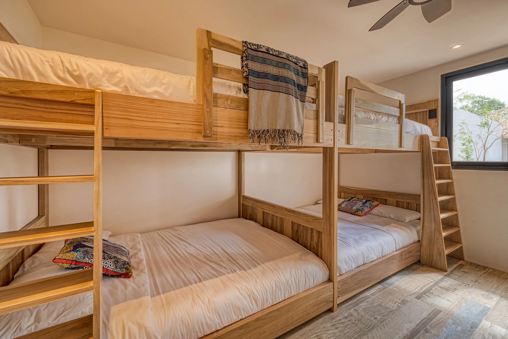 A bedroom with wooden bunk beds, topped with bamboo railings and ladder, near a large window showing some greenery outside.